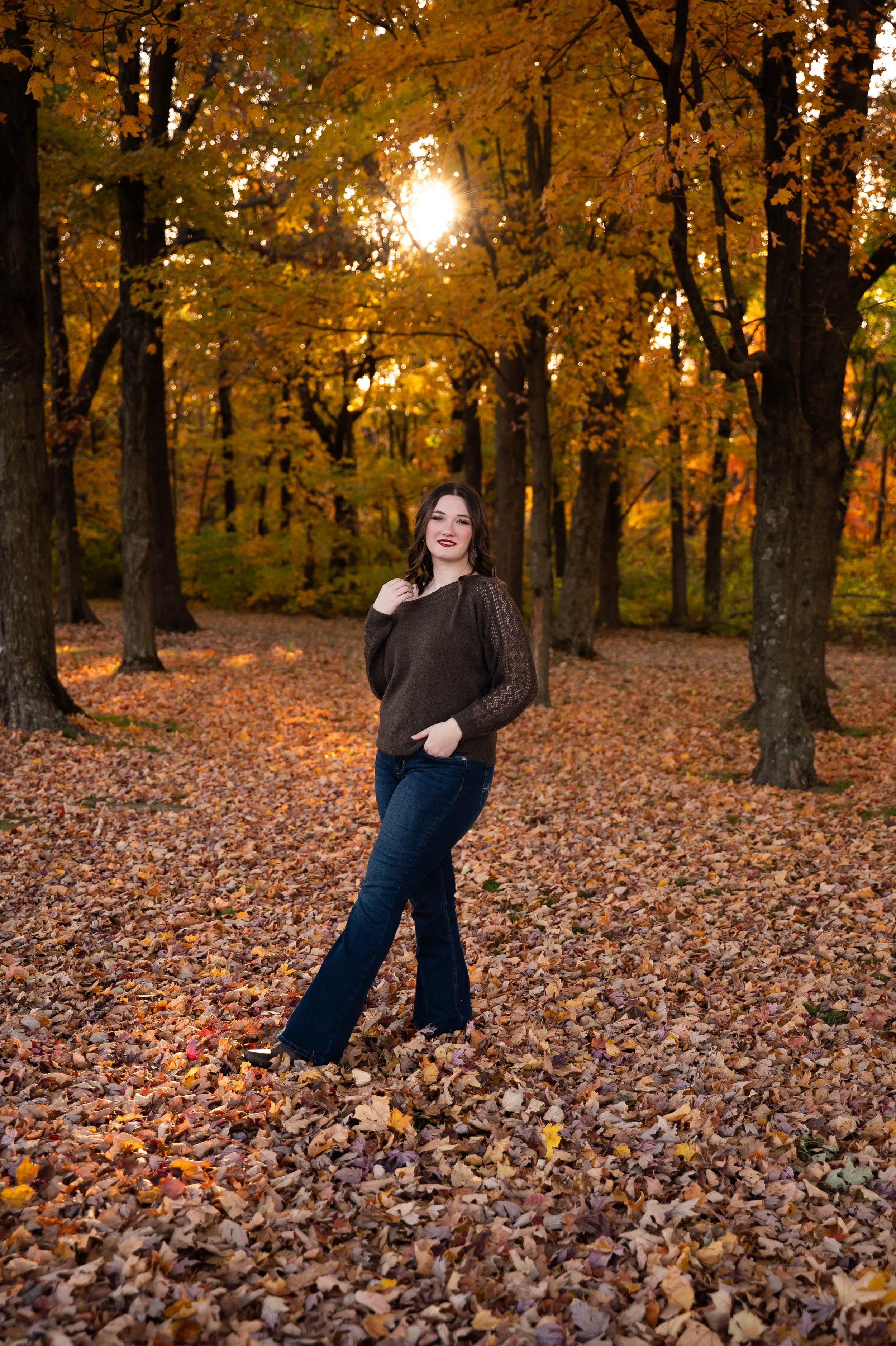 A woman standing in a forest with autumn leaves on the ground and trees with orange and yellow foliage, sun shining through trees in the background.