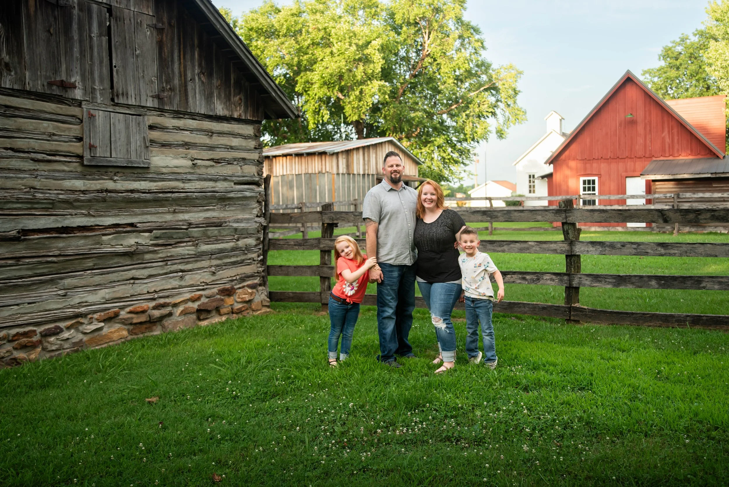 A family of four, consisting of a man, woman, girl, and boy, stands together outdoors on a grassy area near wooden barns and a red house, smiling at the camera.