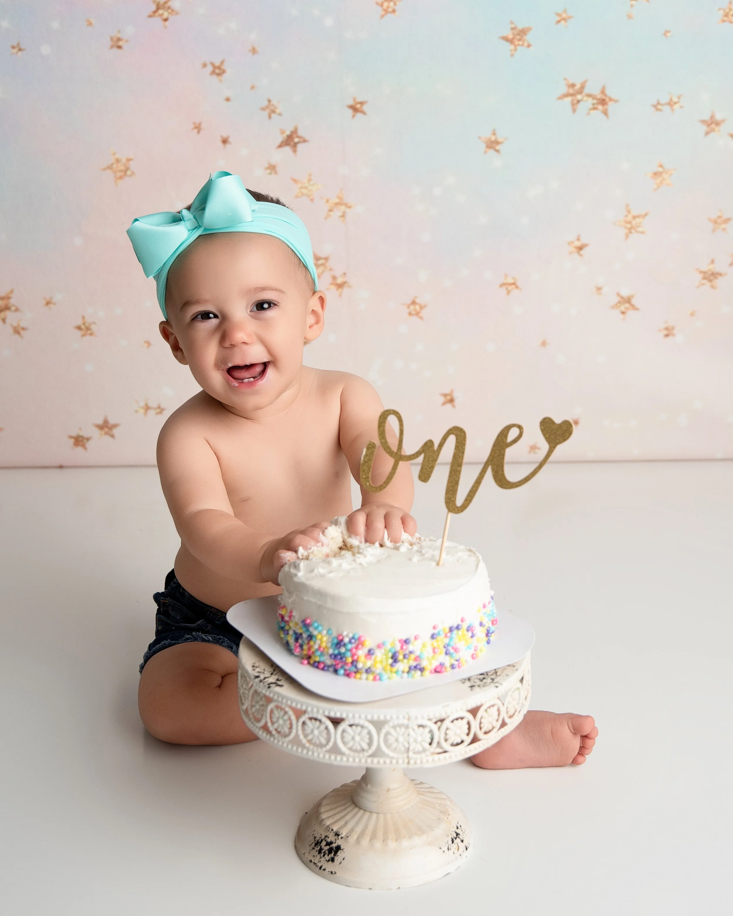 A smiling baby with a blue headband, sitting next to a birthday cake decorated with rainbow sprinkles and a gold 'one' topper, against a starry background.