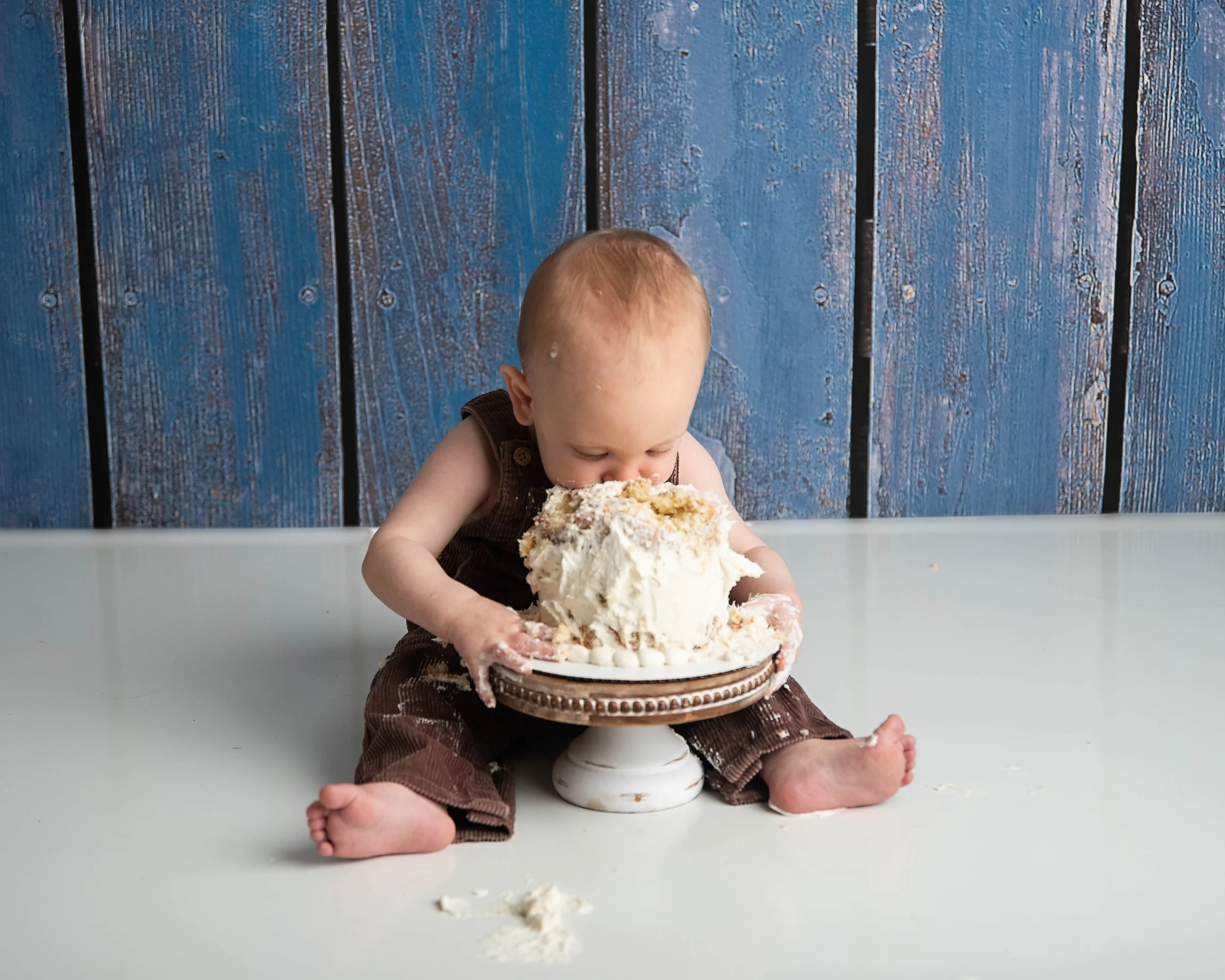 A young baby with light hair sitting on a white surface, eating a large cake with white frosting placed on a pedestal. The baby's face and hands are messy, and the background features a blue wooden wall.