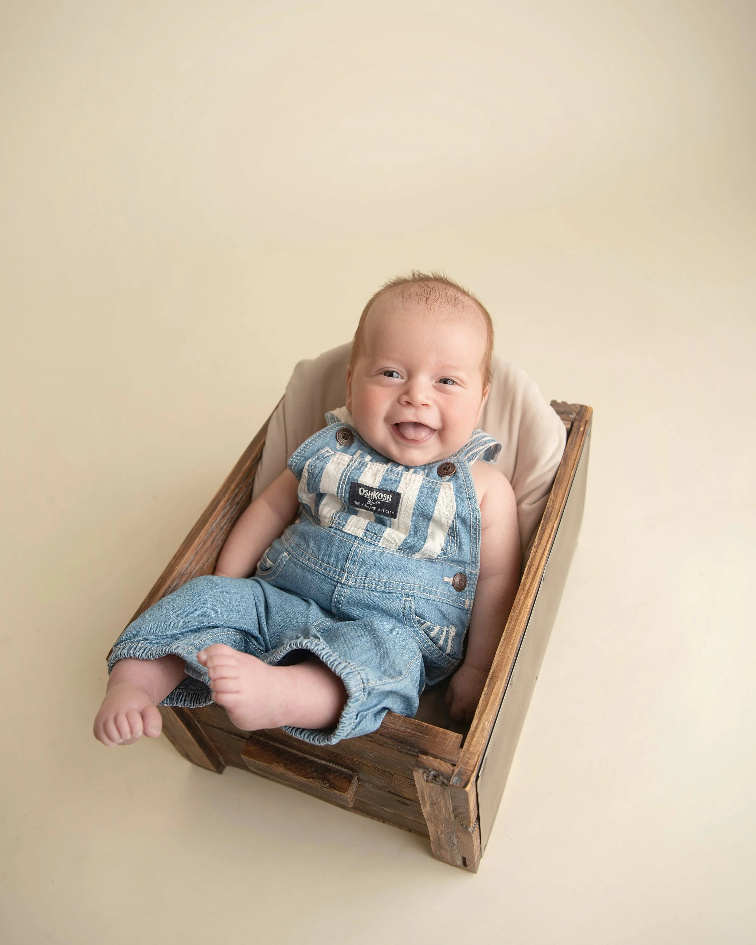 A smiling baby wearing denim overalls, sitting in a wooden box on a plain light-colored background.