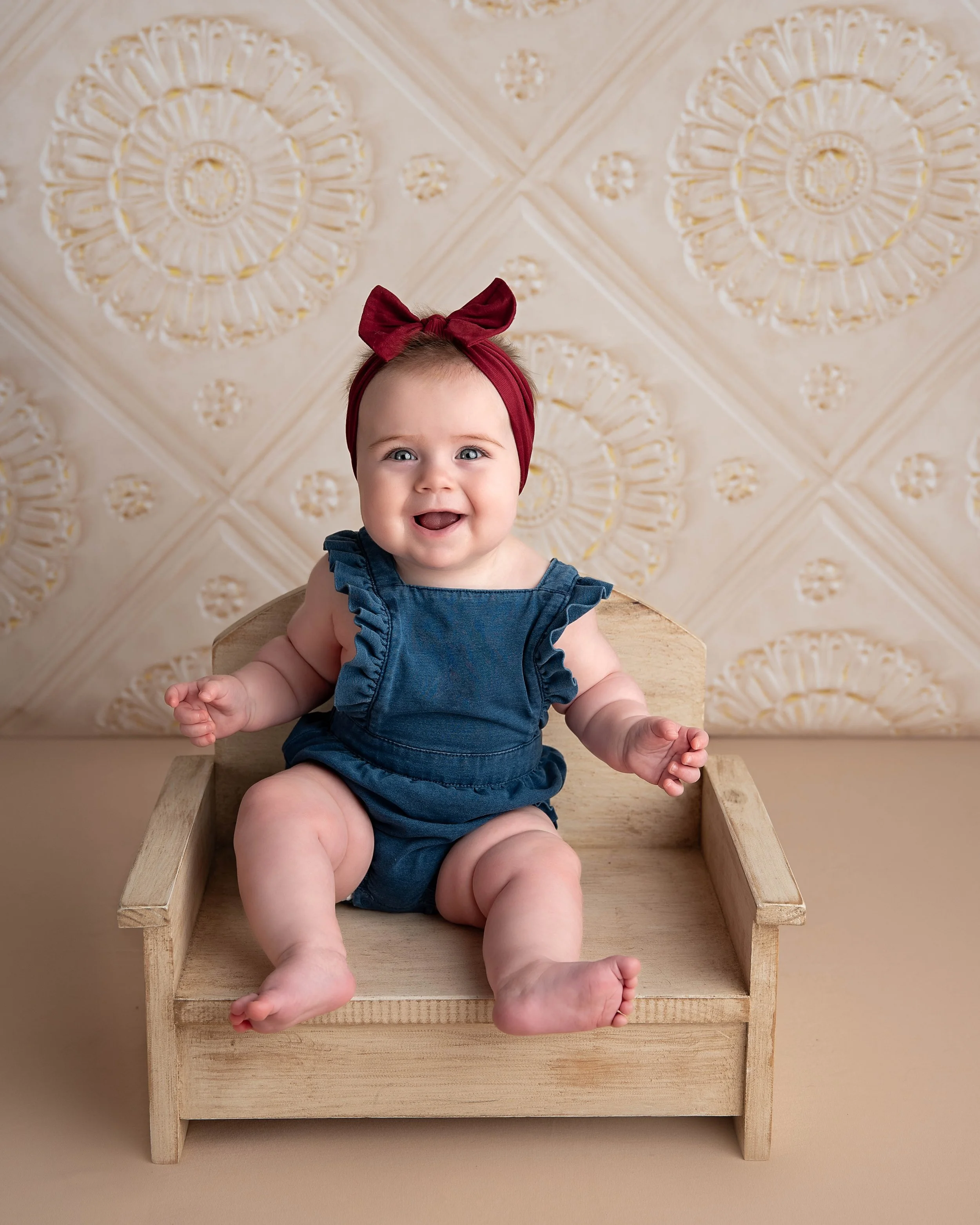A smiling baby girl sitting on a wooden chair, wearing a red headband with a bow and a blue denim outfit, against a beige background with wallpaper featuring floral patterns.