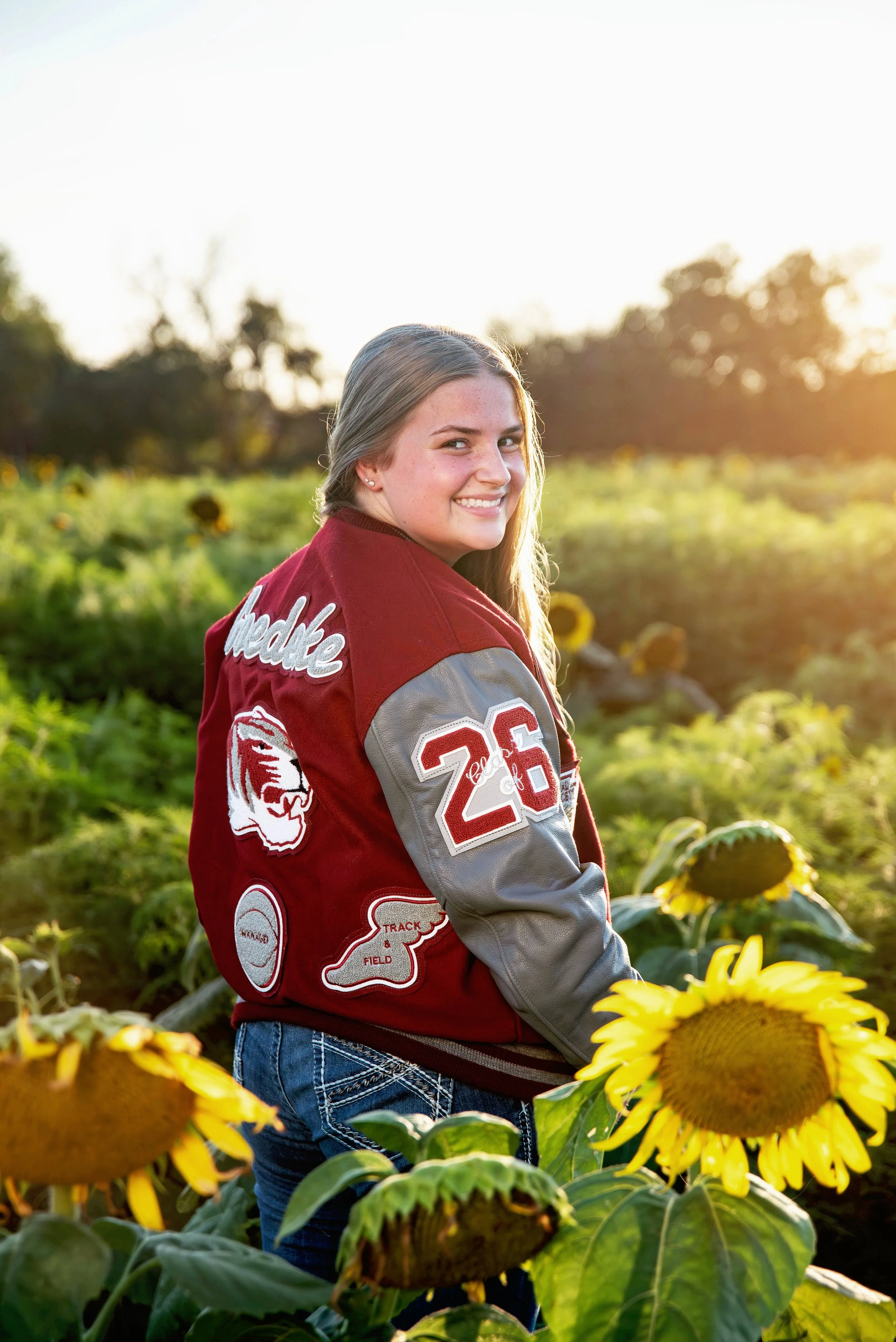 Young woman in a red varsity jacket with patches and the number 26, smiling and standing in a sunflower field during sunset.