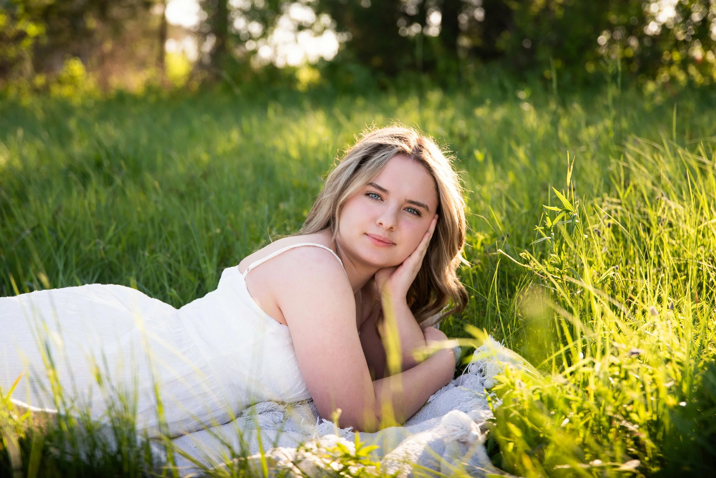 Young woman lying on a blanket in a grassy field, resting her head on her hand, with sunlight and trees in the background.
