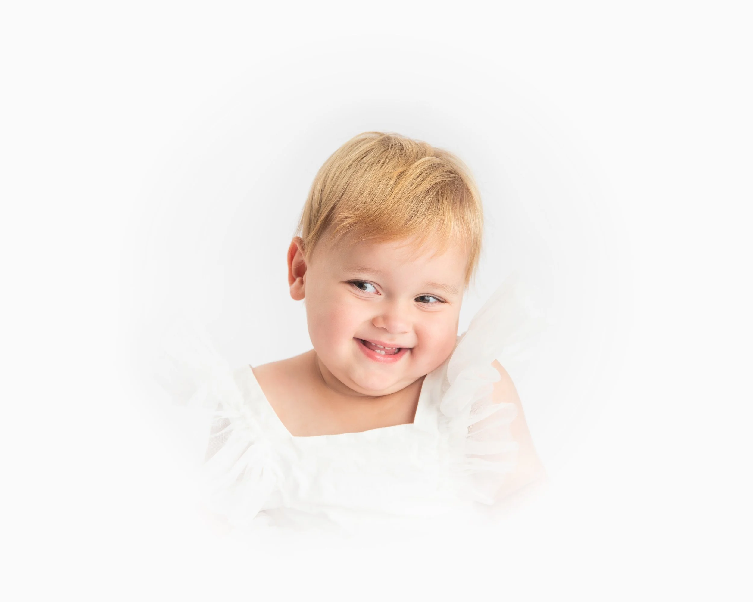 A young child with blonde hair smiling and looking to the side, wearing a white dress with ruffled sleeves, against a white background.