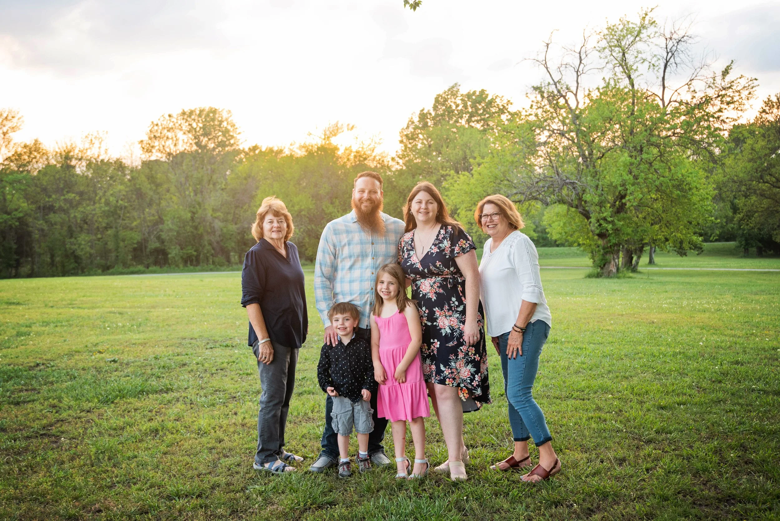 Group of seven people, three adults and three children, standing outdoors in a park during sunset with green grass and trees in the background.