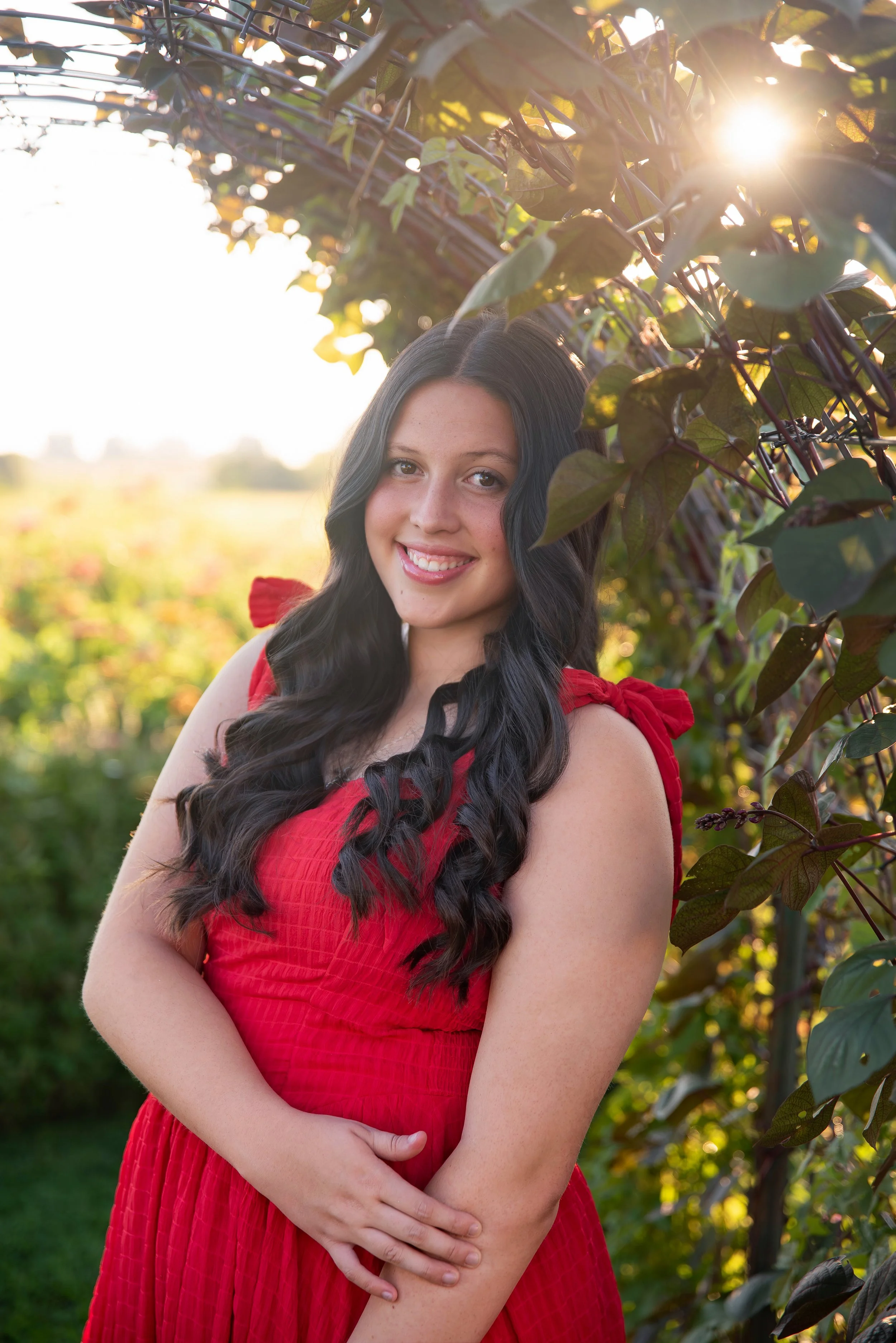 A young woman with long, dark, curly hair, smiling and wearing a red dress with shoulder bows, standing outdoors in a garden or park during golden hour with the sun shining through the leaves.