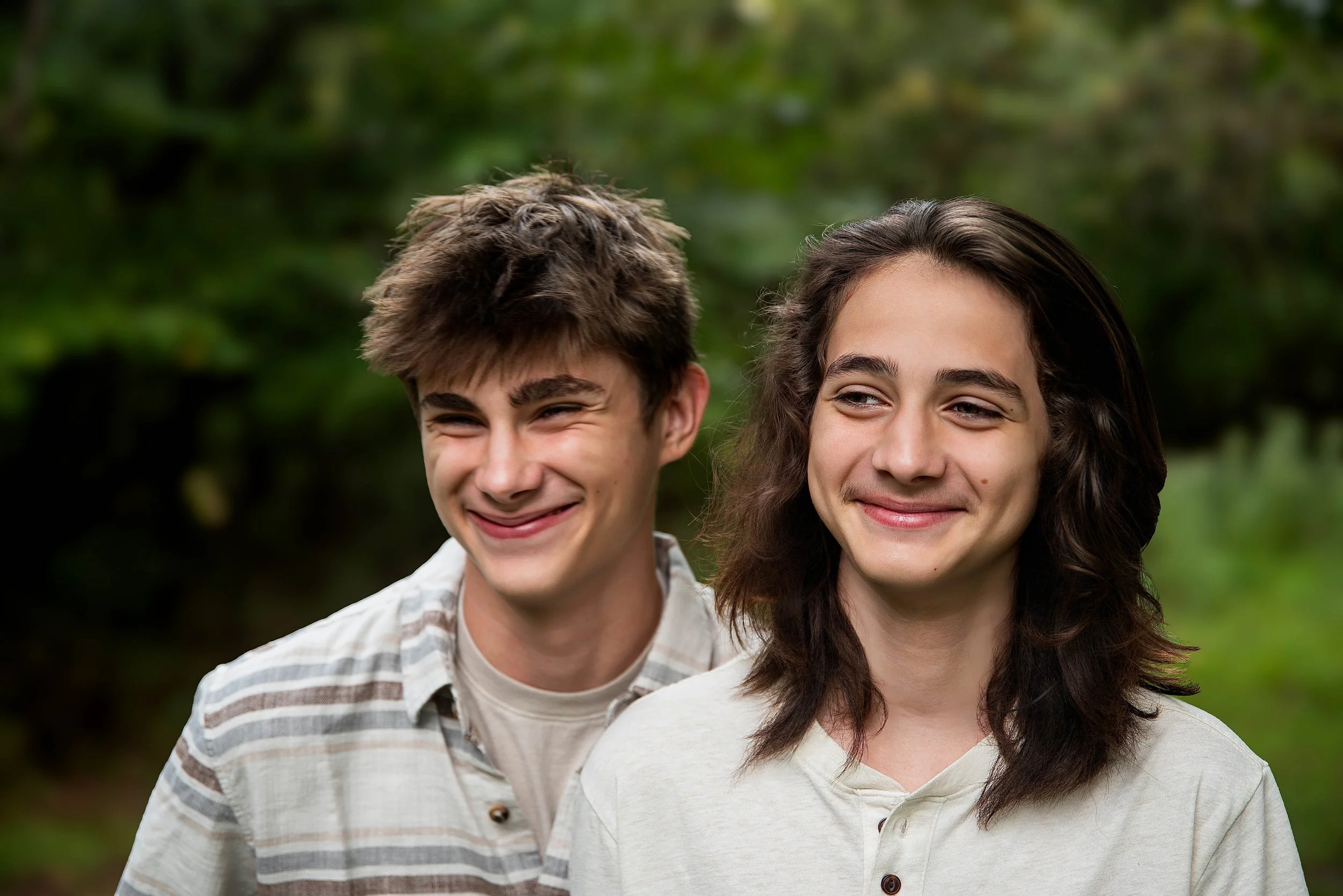 Two young men smiling and standing outdoors in a green, wooded area.