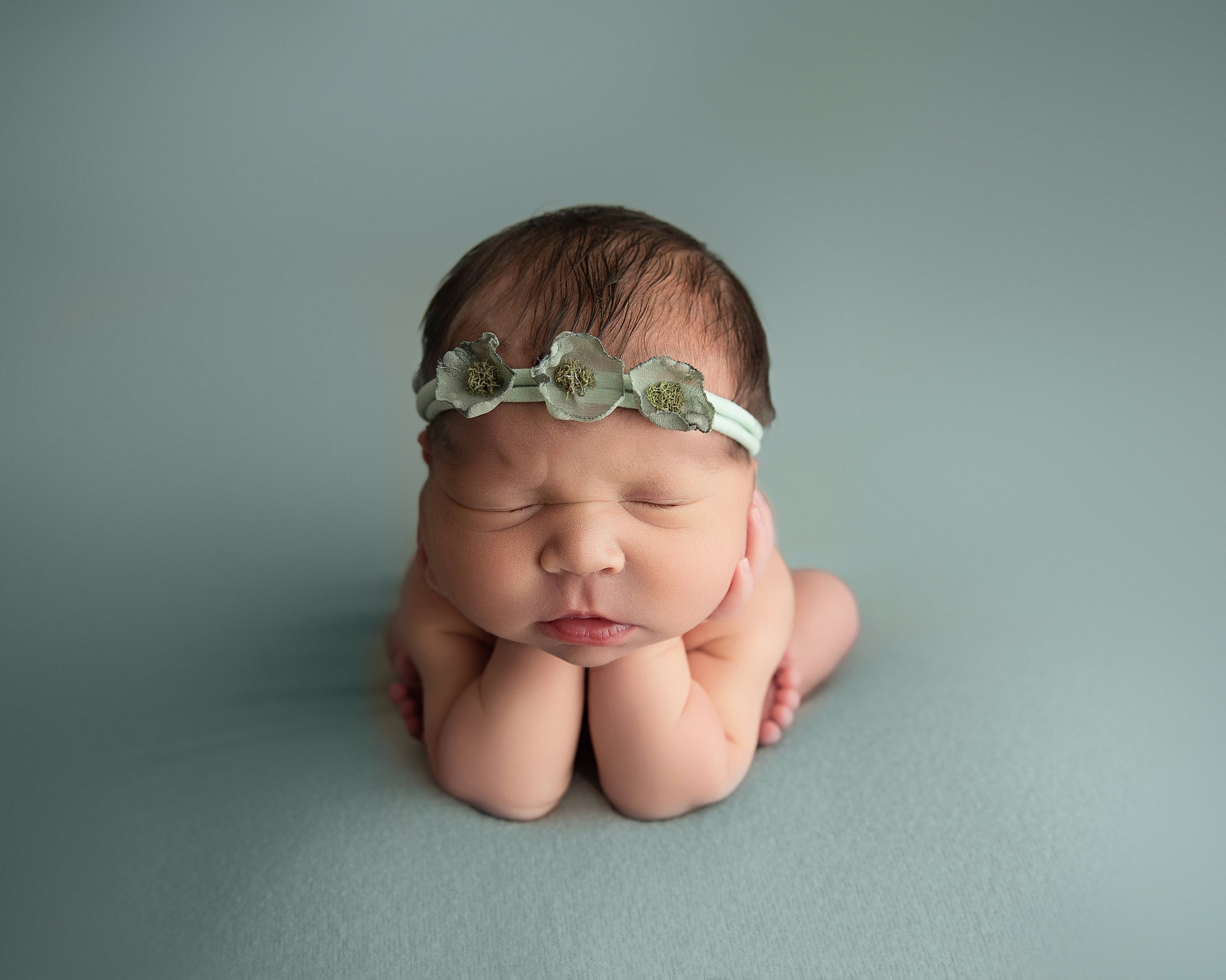 A sleeping newborn baby with a floral headband, lying on its tummy with hands under chin, on a light blue surface.