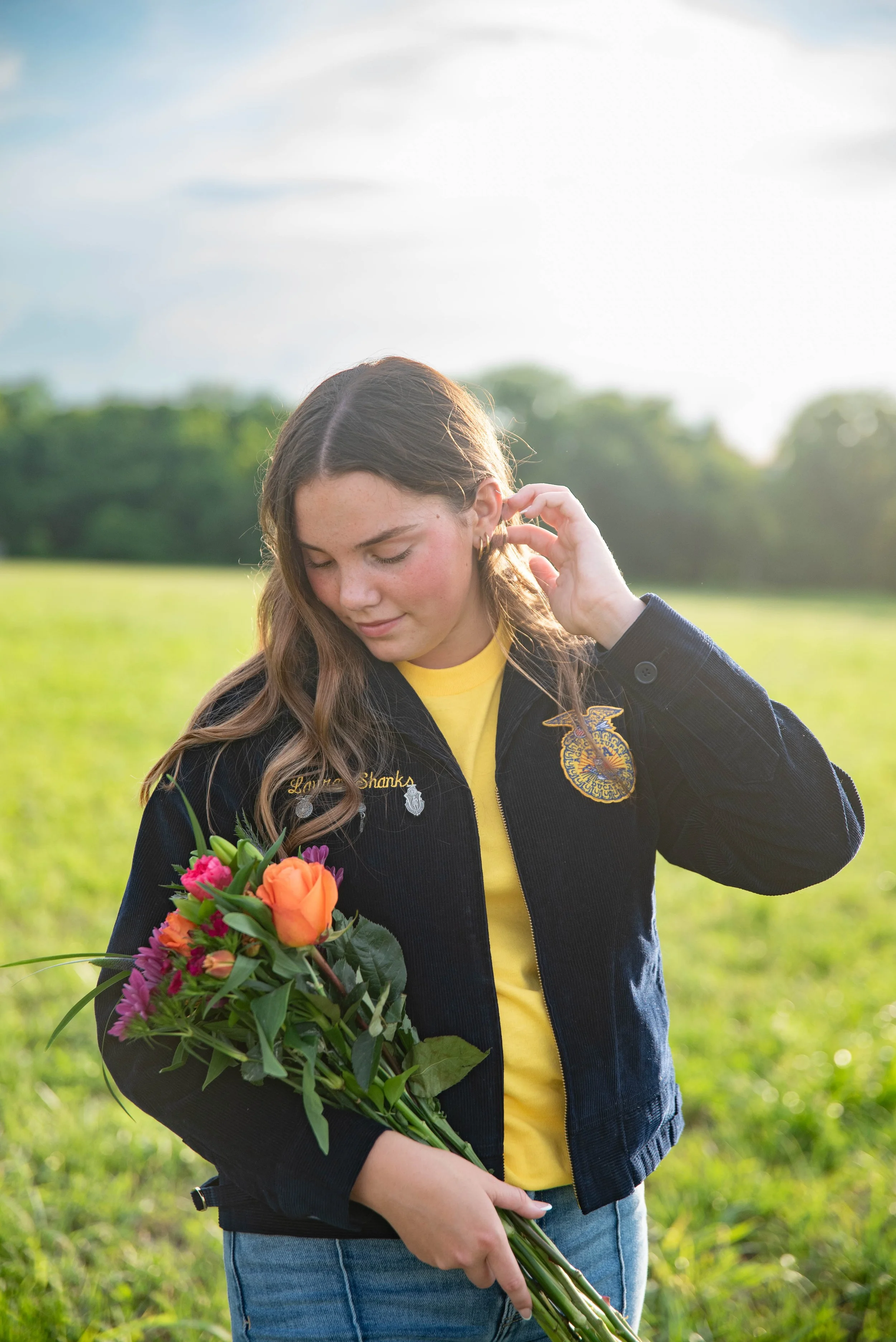 A young woman holding a bouquet of colorful flowers, standing outdoors in a green field with trees in the background, wearing a dark jacket and smiling gently.
