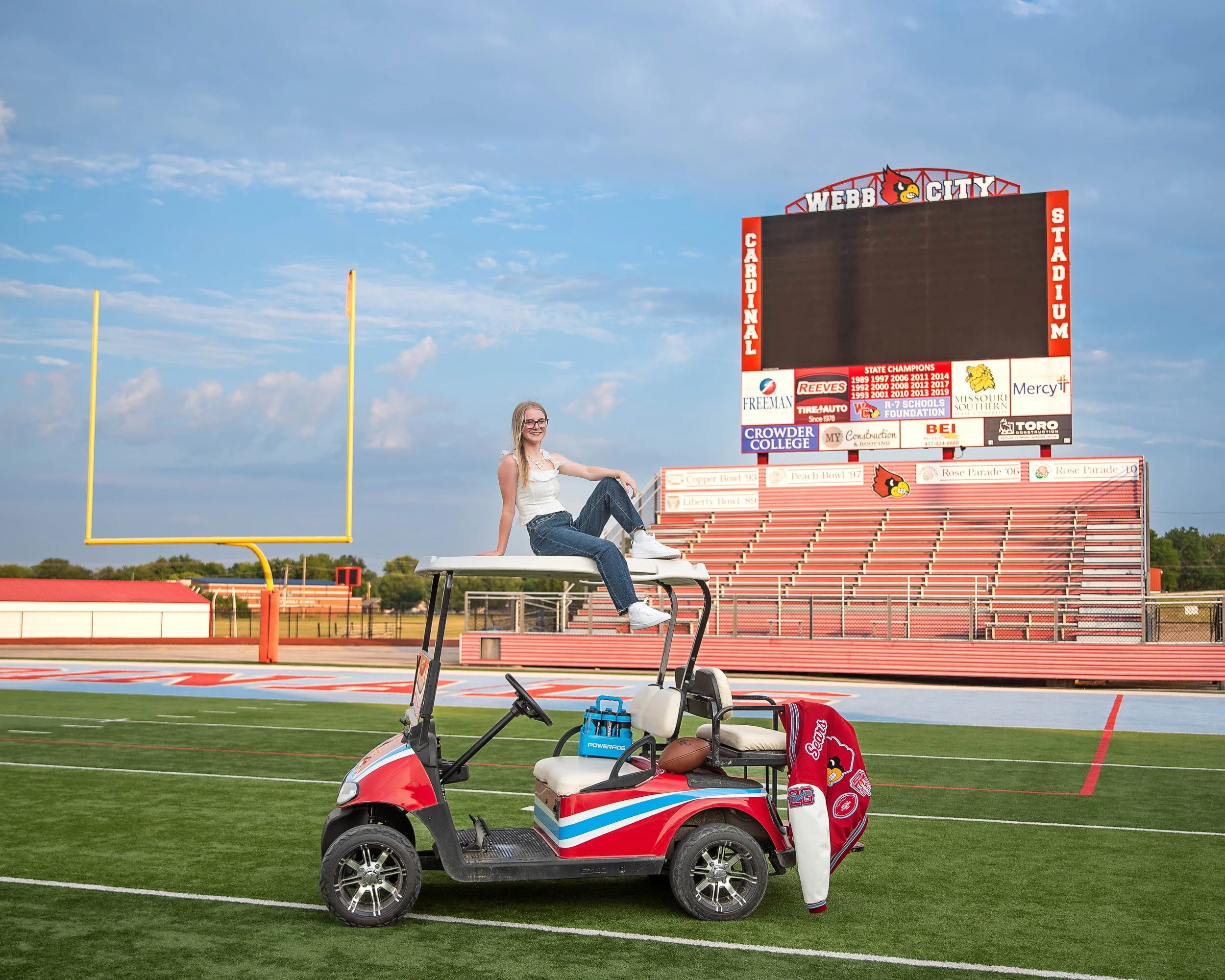 A young woman sits on top of a golf cart on a football field. She is smiling and wearing glasses, a white tank top, and jeans. The cart has a red, white, and blue design and holds a golf club bag and a water bottle. In the background, there are stadi