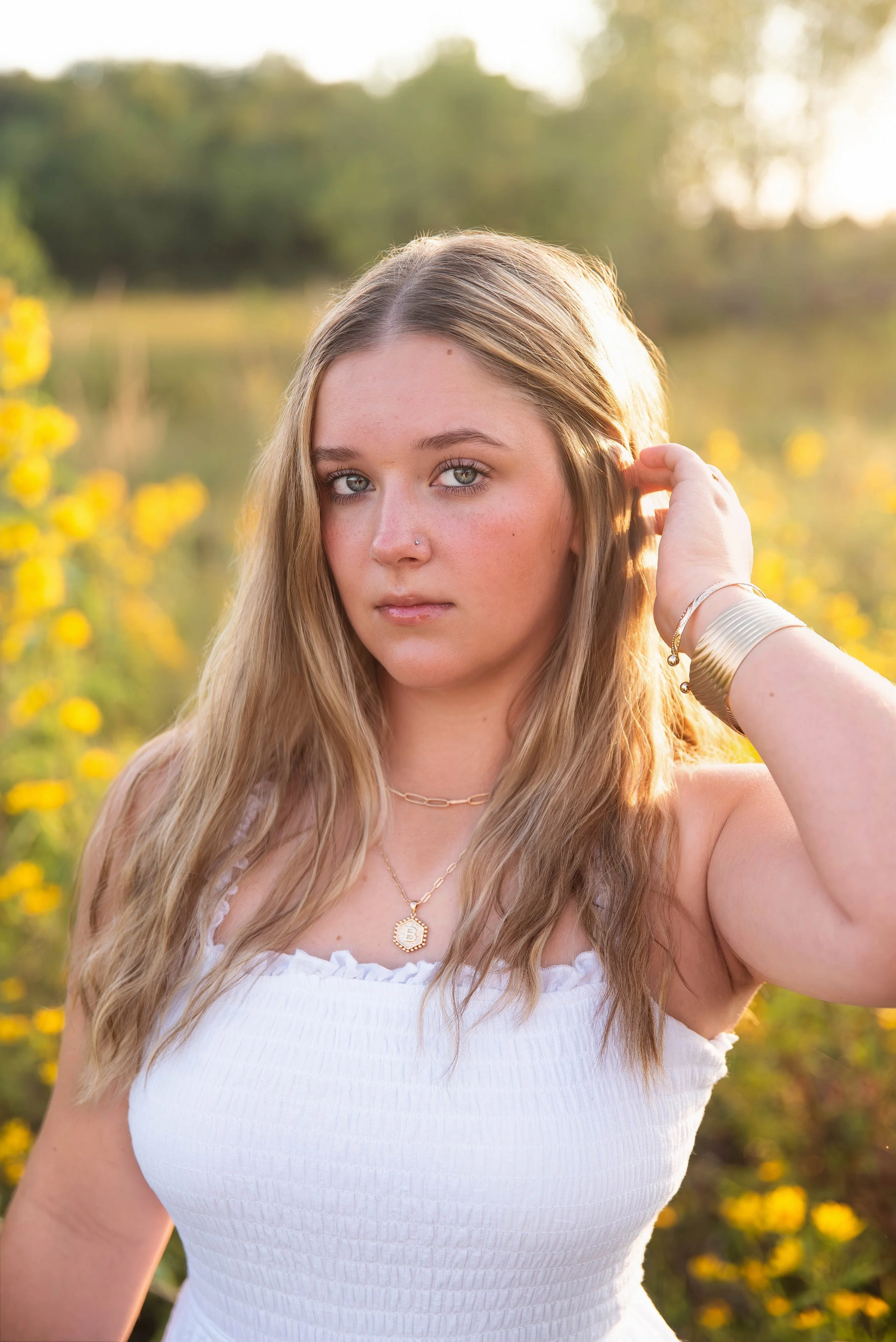 A young woman with long blonde hair and blue eyes stands outdoors during sunset, wearing a white sleeveless top, gold jewelry, and placing her hand behind her ear amidst a background of yellow flowers and greenery.