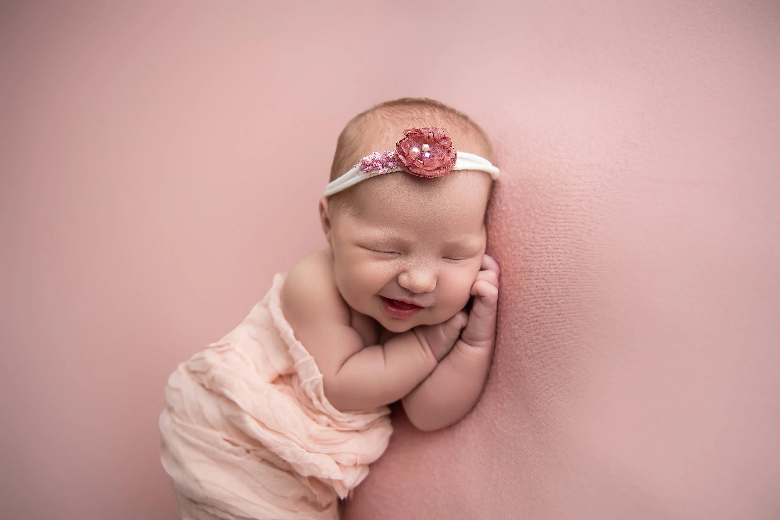 Smiling sleeping newborn baby girl lying on pink blanket with a pink headband with fabric flower and pearls, on a pink background.
