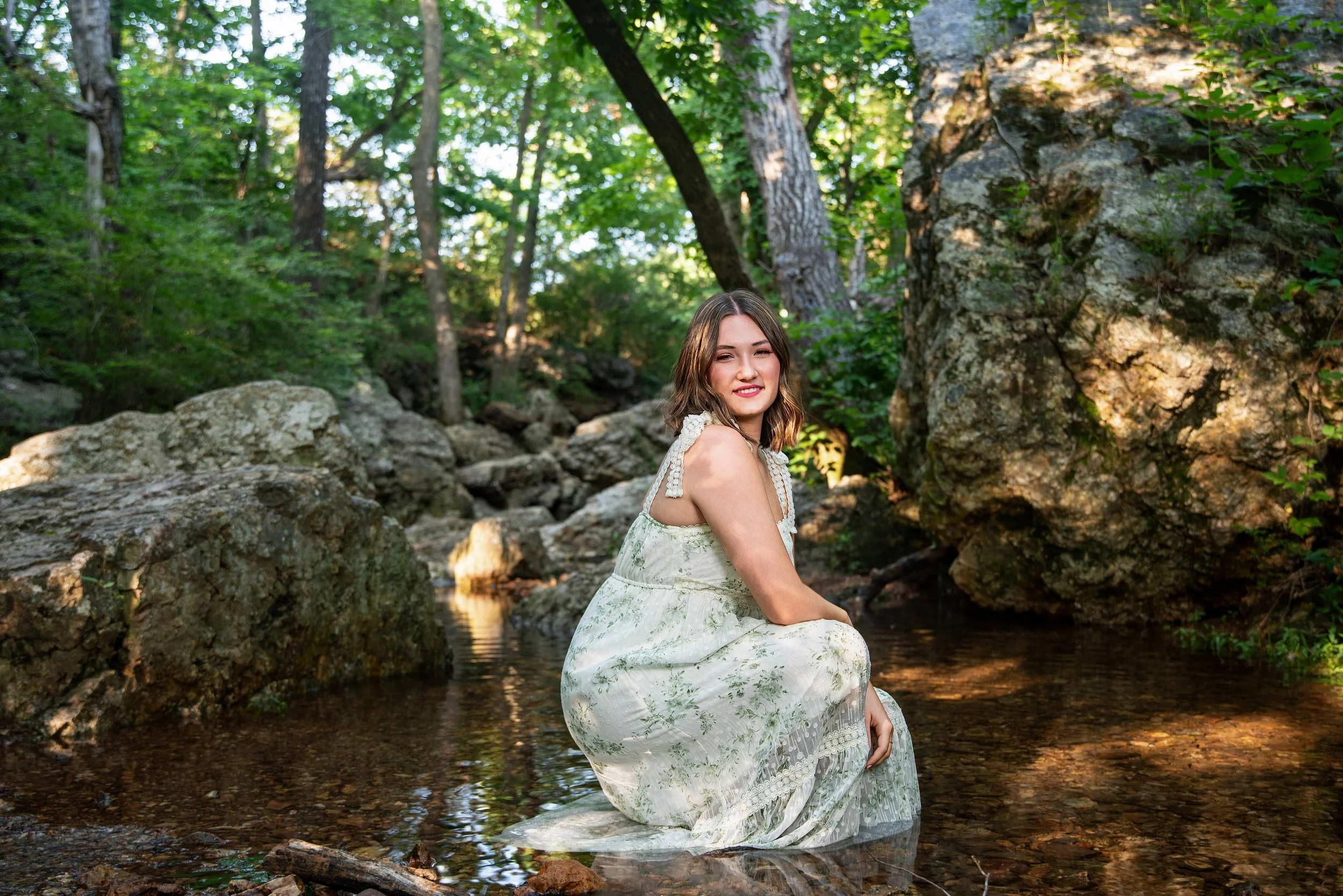 A young woman in a white floral dress kneeling in a creek surrounded by rocks and lush green trees.