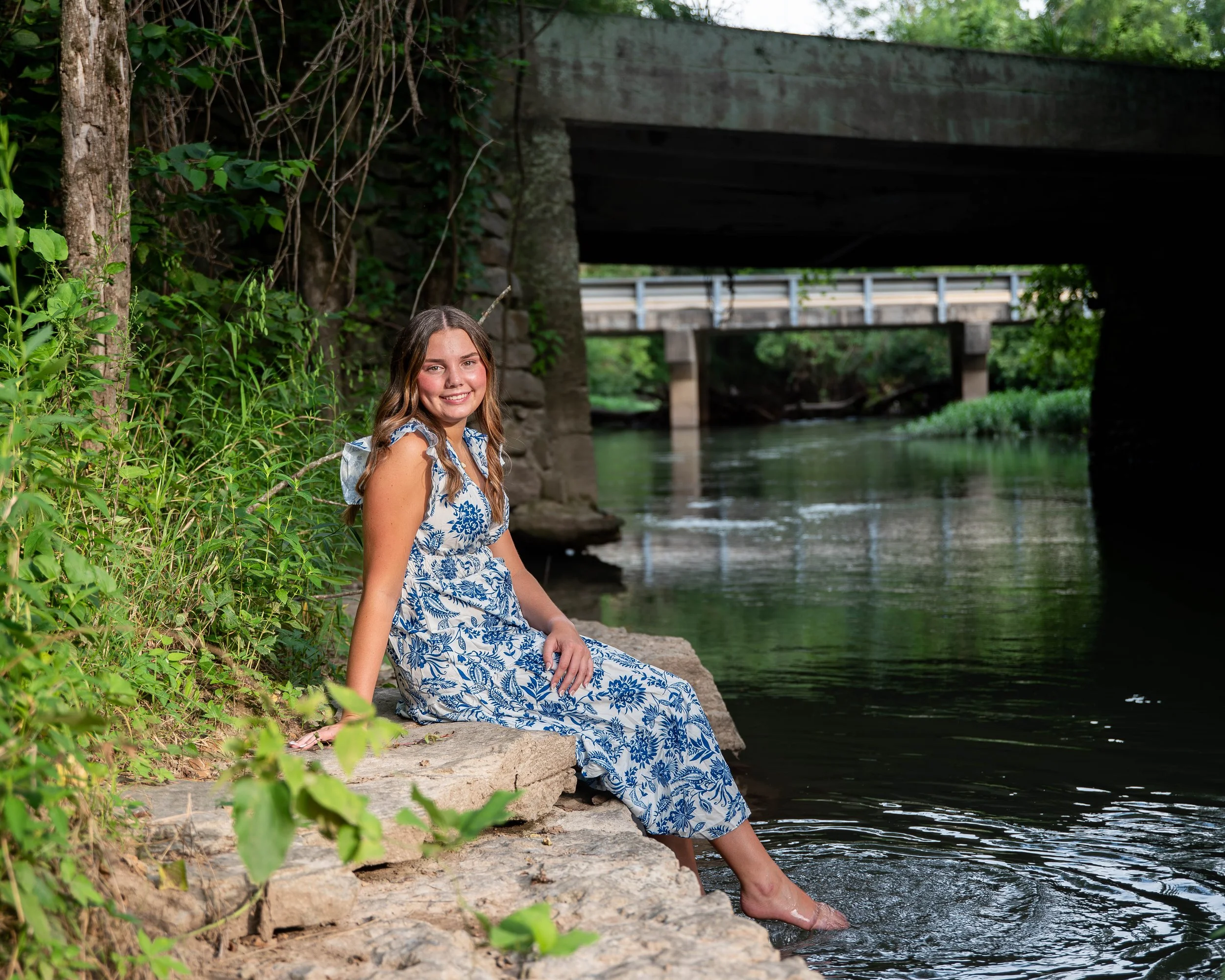 A woman in a blue and white floral dress sitting on rocks by a river with greenery and fallen leaves, with a bridge overhead in a natural outdoor setting.
