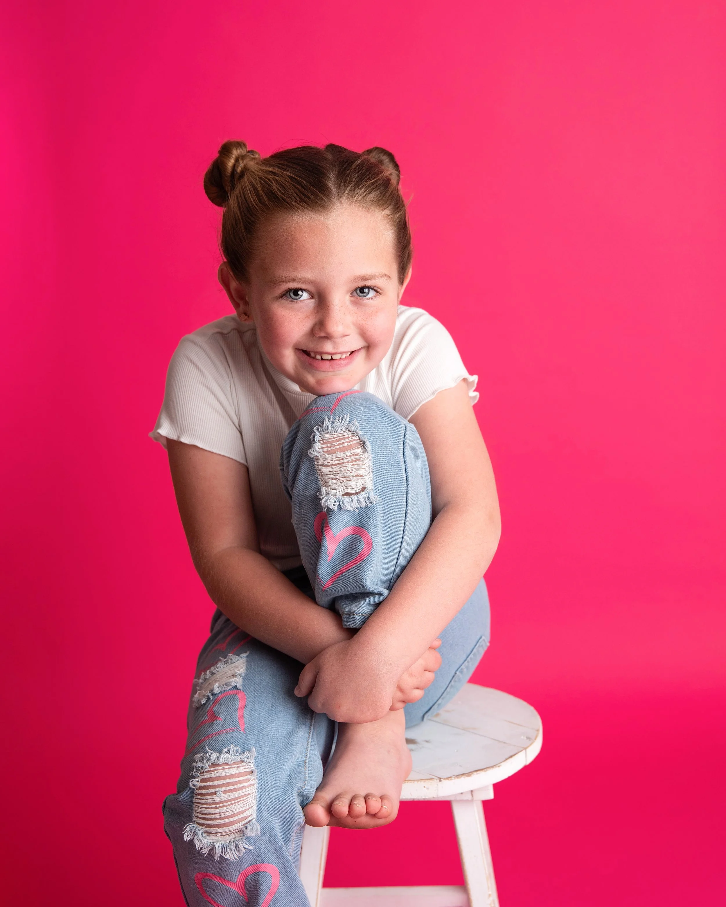 A smiling young girl with braided hair, wearing a white t-shirt and ripped jeans with pink heart designs, sitting on a white stool against a bright pink background.