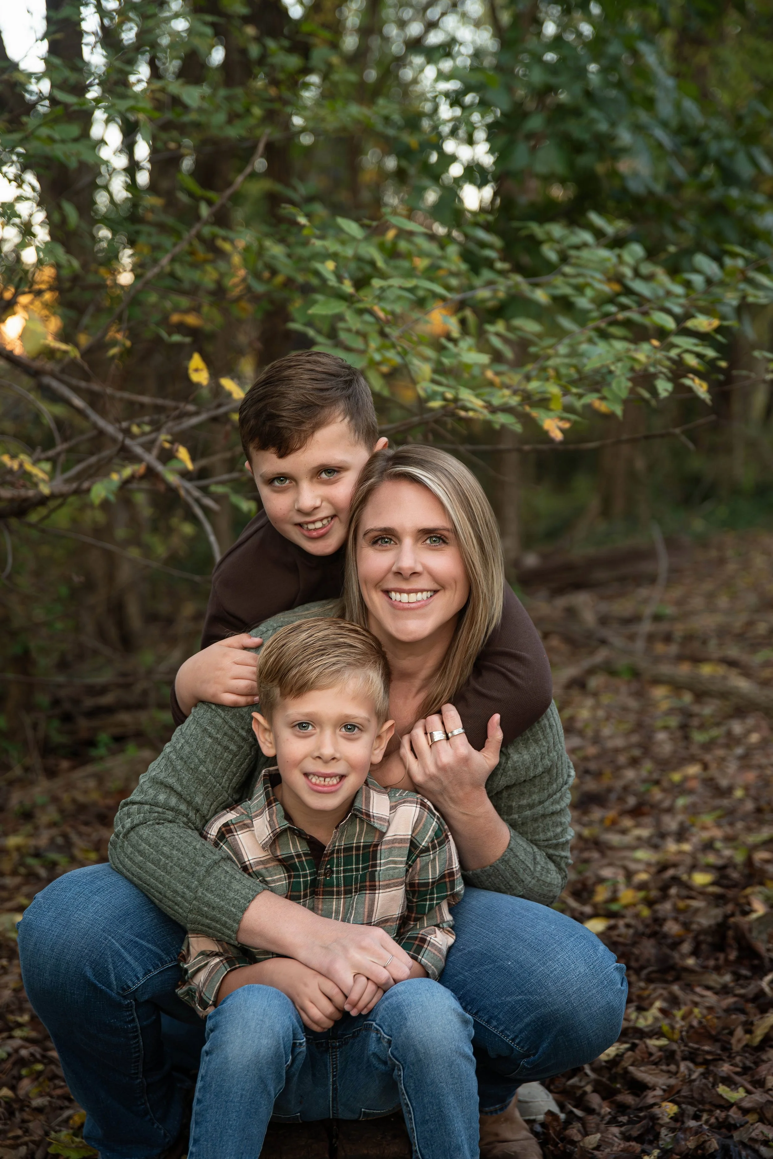 A woman with two young boys outdoors in a wooded area, smiling at the camera, showing a close family portrait.