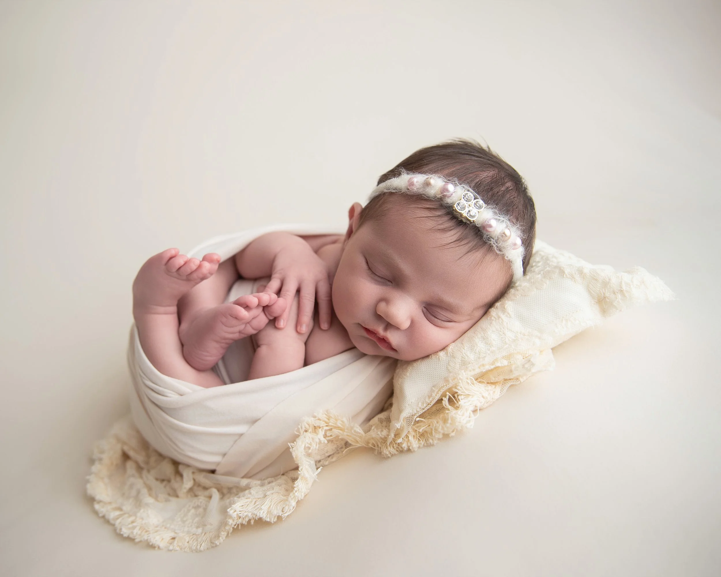 A peacefully sleeping newborn baby wrapped in a white cloth, laying on a soft cream-colored blanket with a textured edge, wearing a pearl and rhinestone headband.