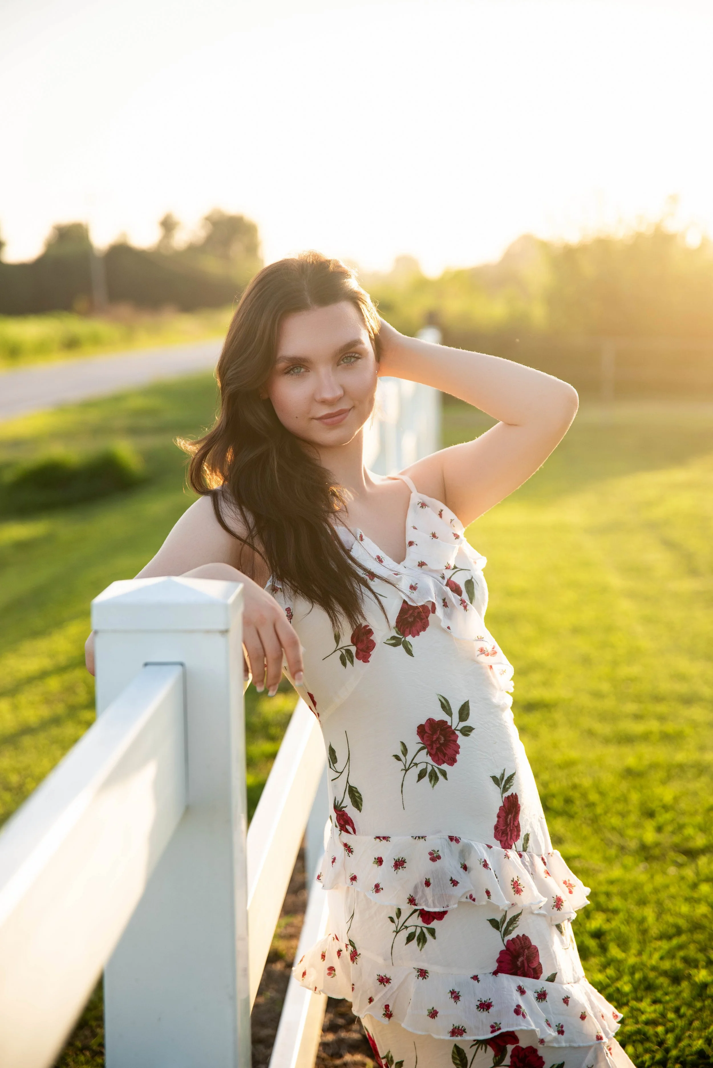 A young woman with long dark hair standing outdoors during sunset, leaning on a white fence with a green field in the background, wearing a white dress with red floral patterns.