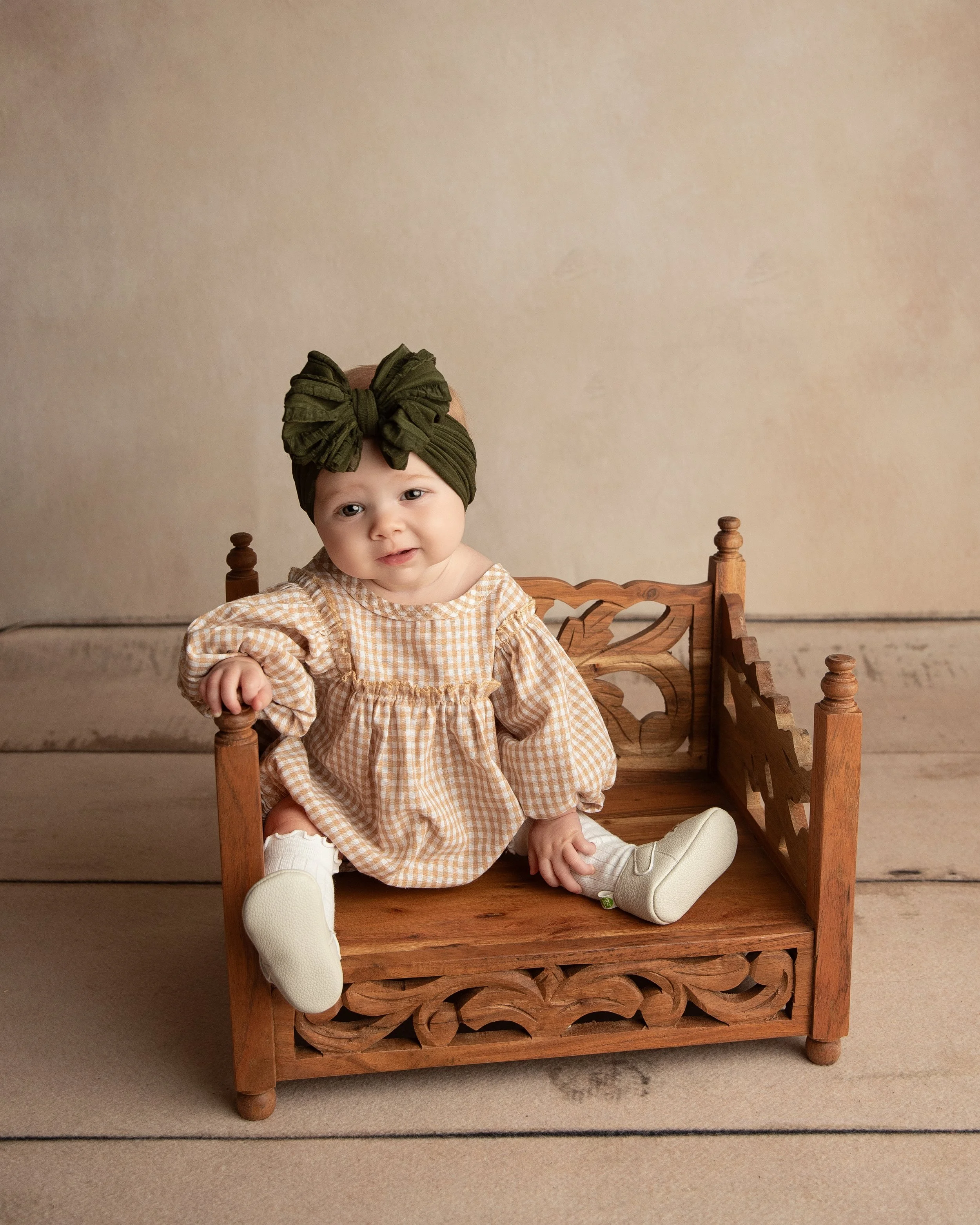 A baby sitting on a wooden miniature bed, wearing a brown checkered dress, white socks, and beige shoes, with a large green headwrap, against a plain beige background.