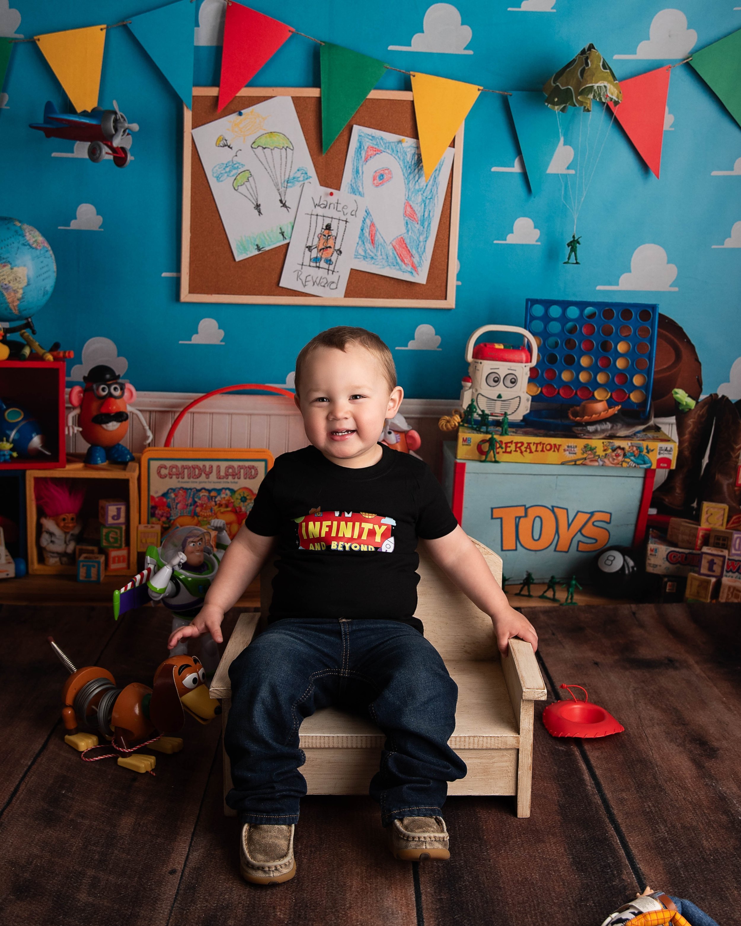 A young boy sitting on a small wooden chair in a playroom filled with toys, with a bright blue wall decorated with clouds and drawings of parachutes and a wanted poster. There are colorful banners hanging from the ceiling and various toys around him.