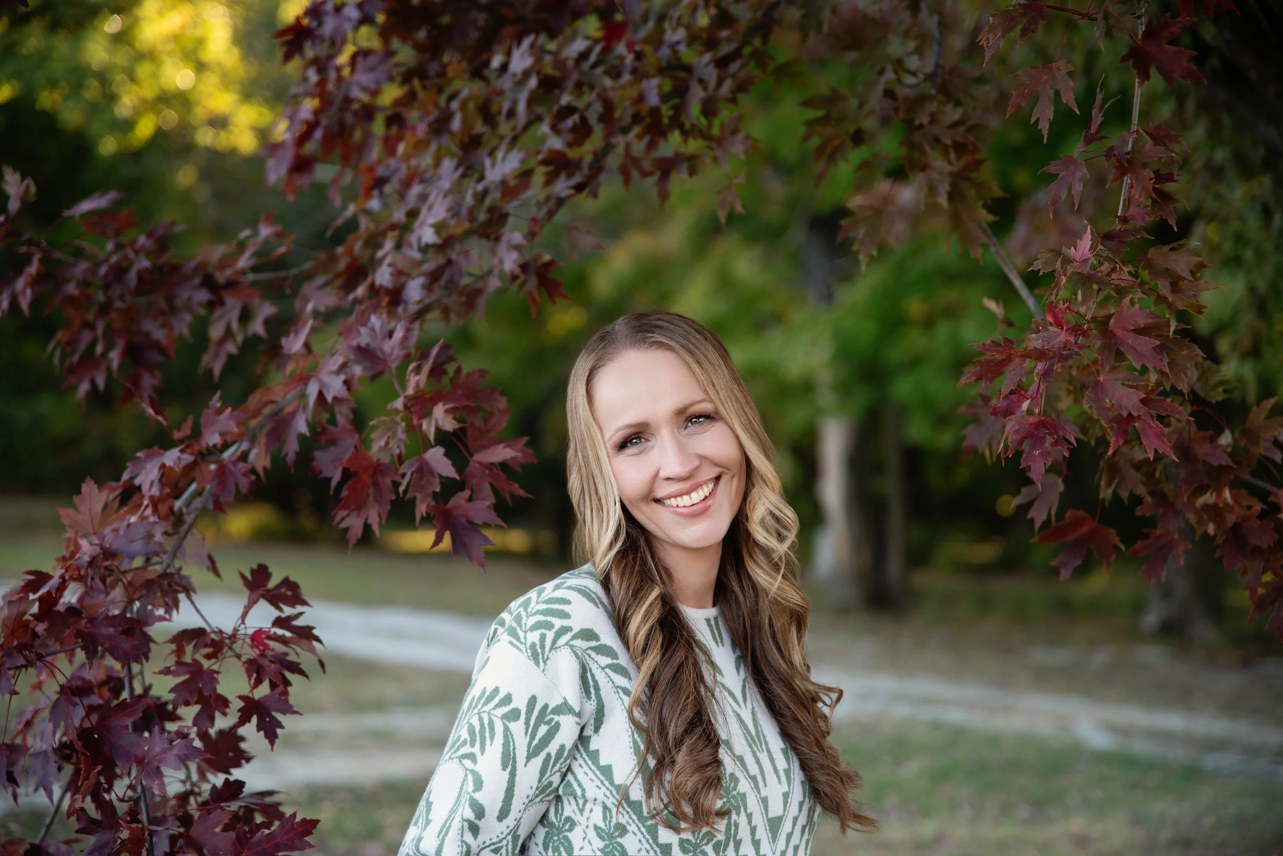A smiling woman with long wavy blonde hair wearing a white and green patterned shirt outdoors in front of red and green foliage.