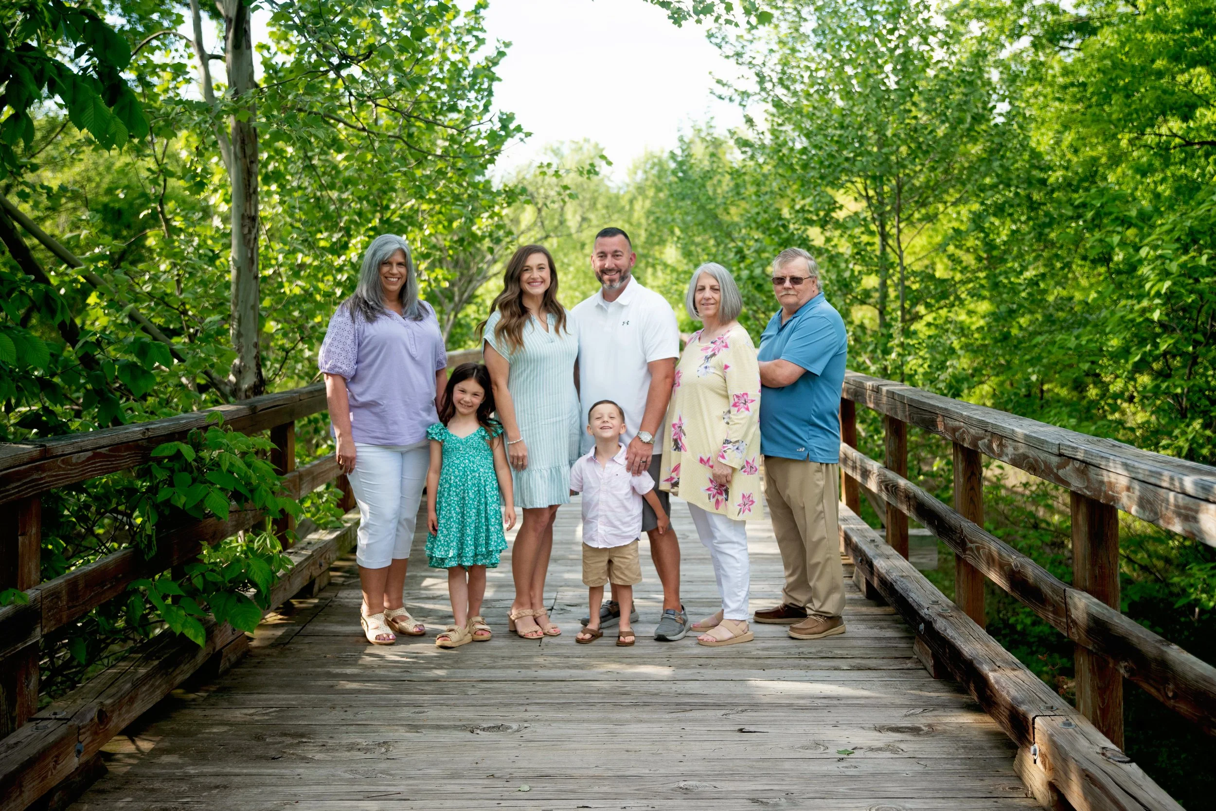 A multi-generational family standing on a wooden bridge in a lush green forest, smiling at the camera.