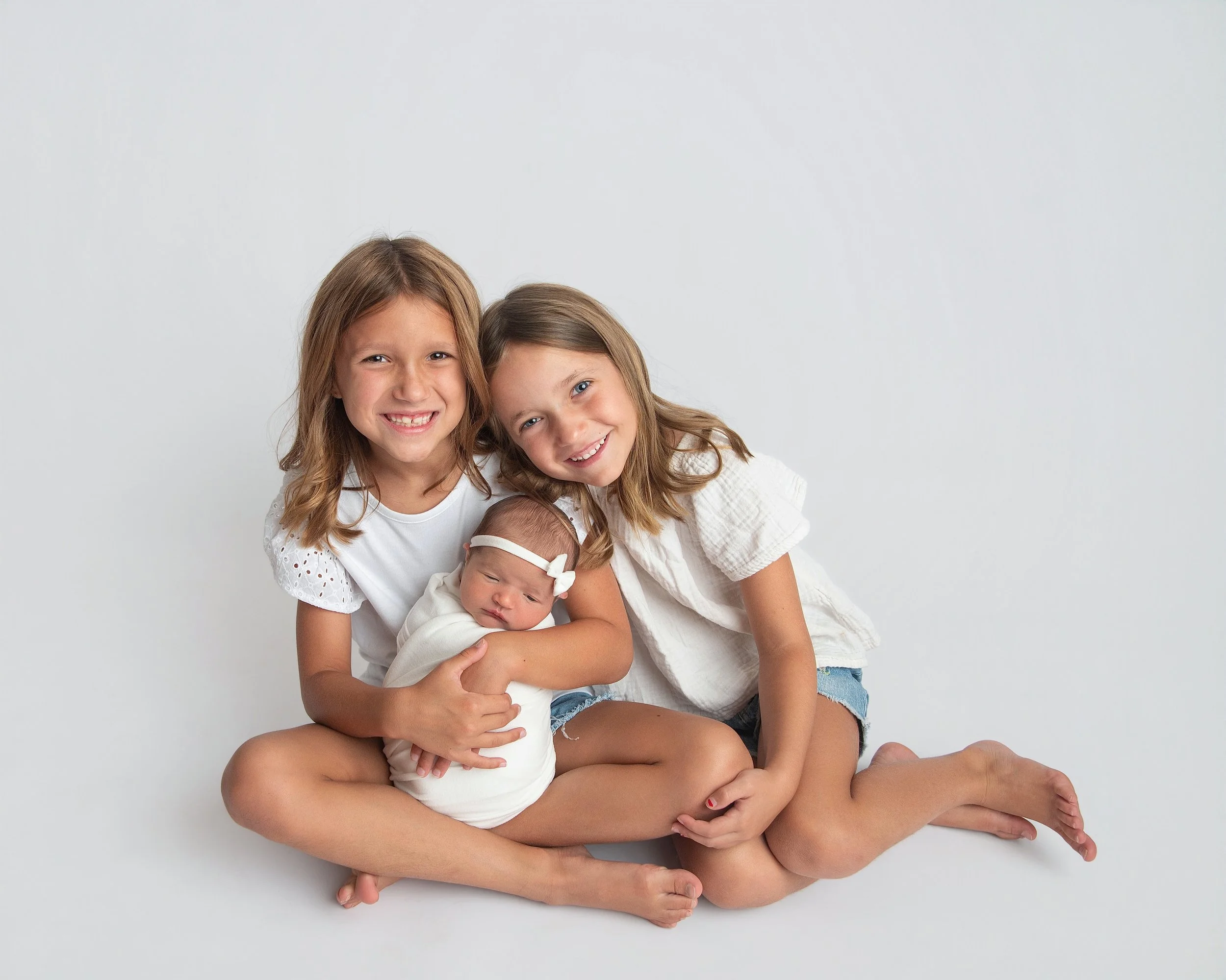 Two young girls and a newborn baby girl pose for a photo against a plain white background. The older girls are smiling, with one holding the baby, who is dressed in a white outfit and bow headband.