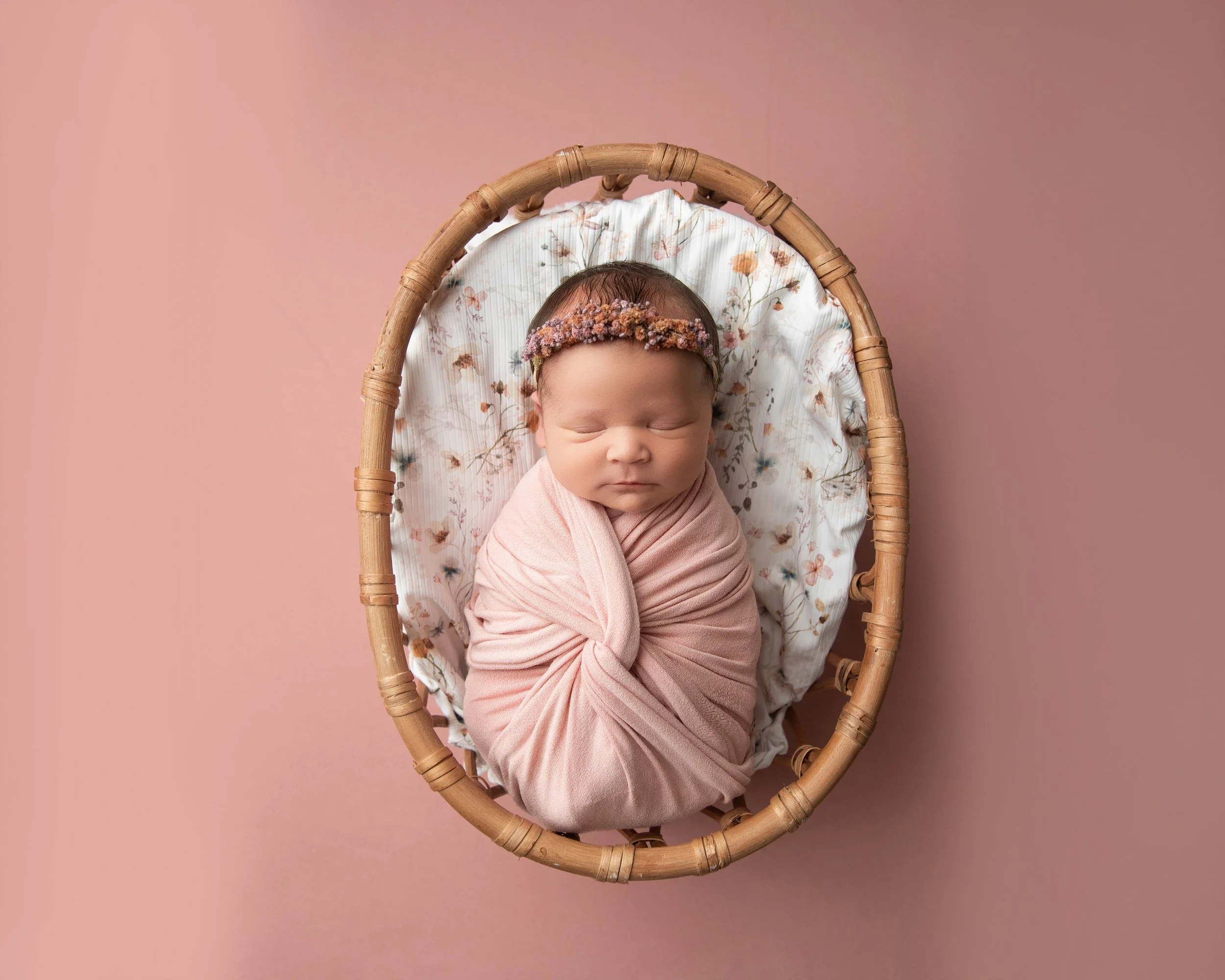 A sleeping baby girl wrapped in a pink blanket, lying in a wicker basket with a floral blanket inside, on a pink background.