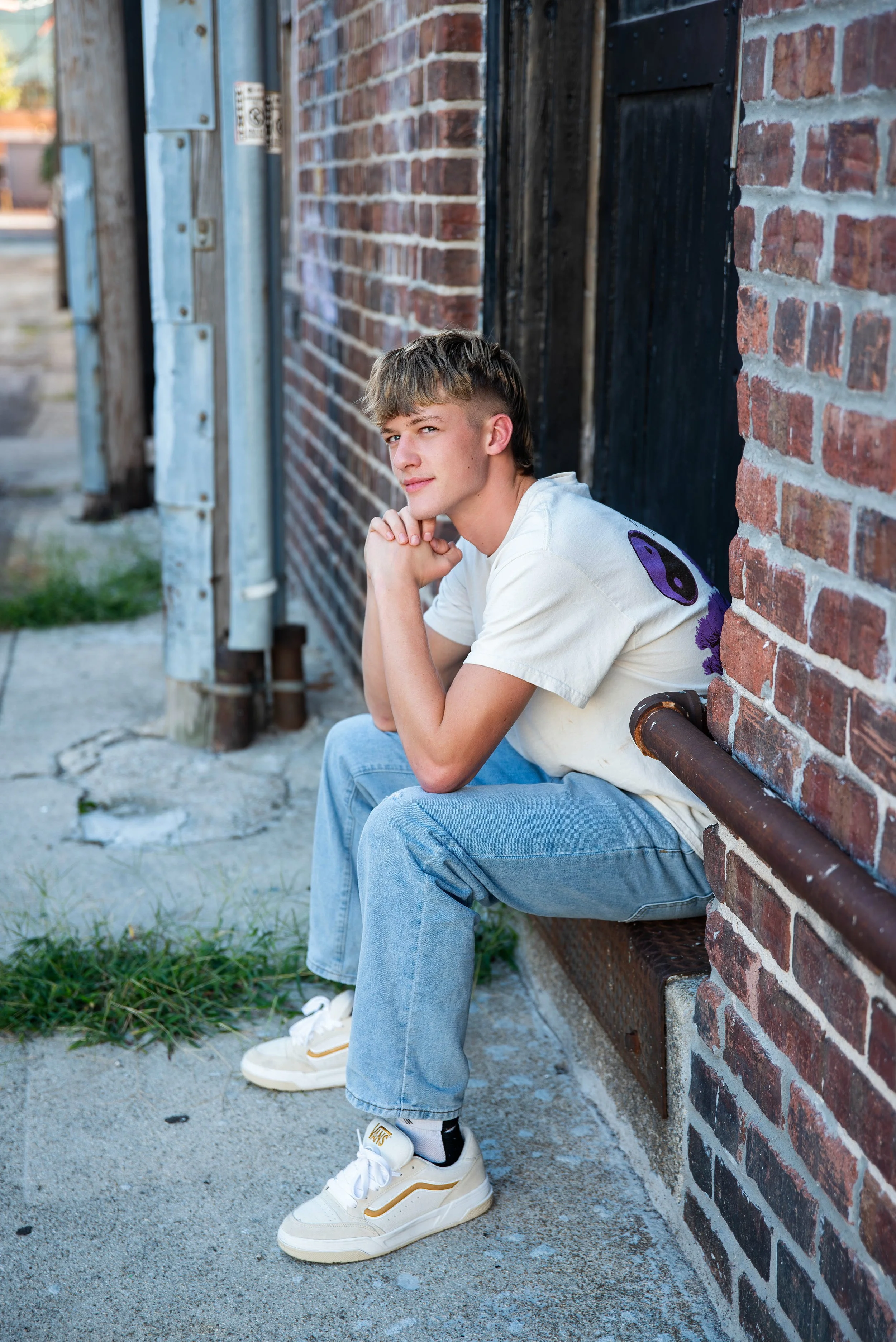 A young man with short, light brown hair and a slight smile, sitting on a ledge outside against a brick wall, wearing a white T-shirt, light blue jeans, and white sneakers. He has his chin resting on clasped hands and is looking at the camera.