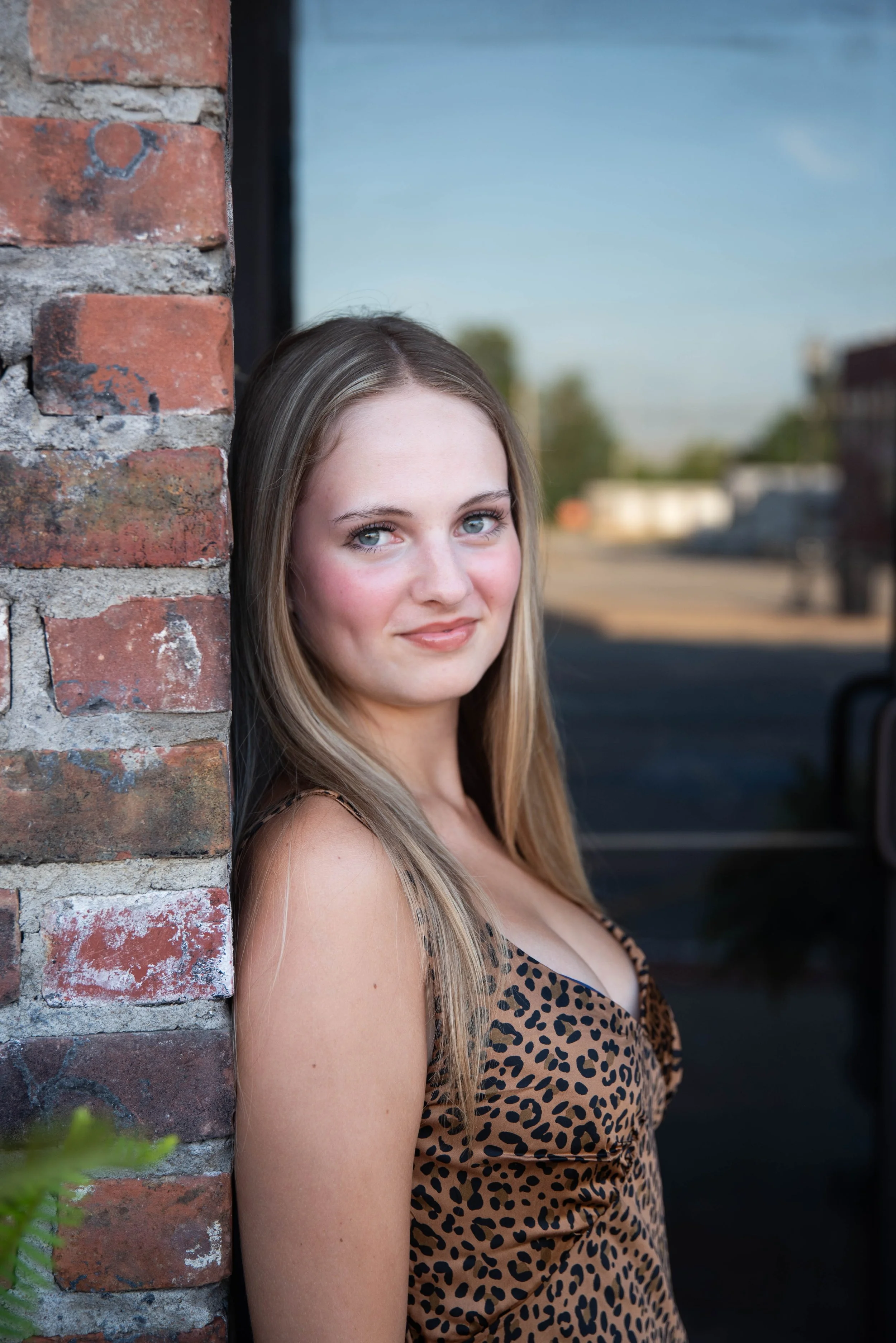 Young woman with long blonde hair and blue eyes leaning against a brick wall outdoors, wearing a leopard print top.