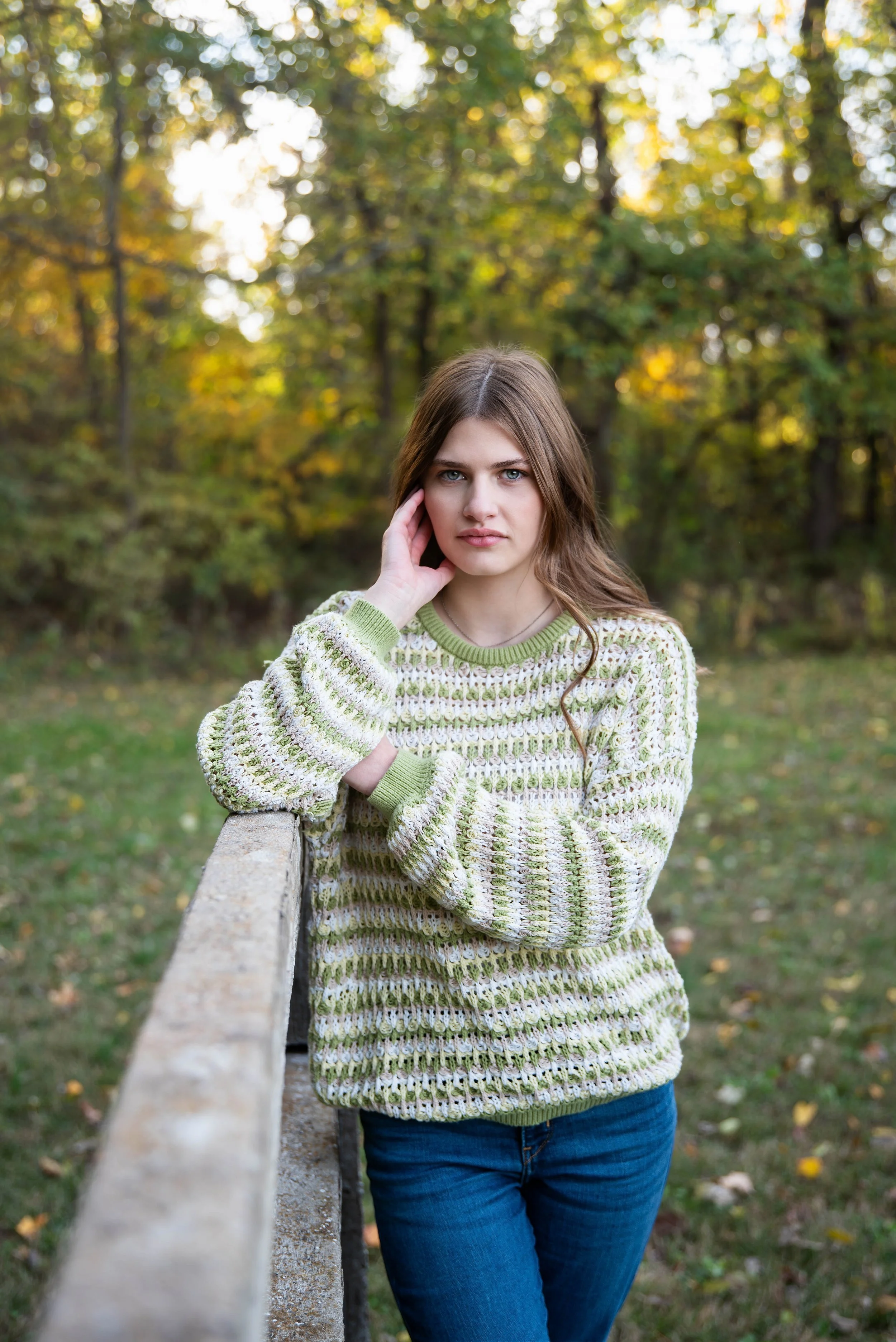 A young woman with long brown hair and blue eyes leaning on a wooden fence in a park with autumn foliage in the background.