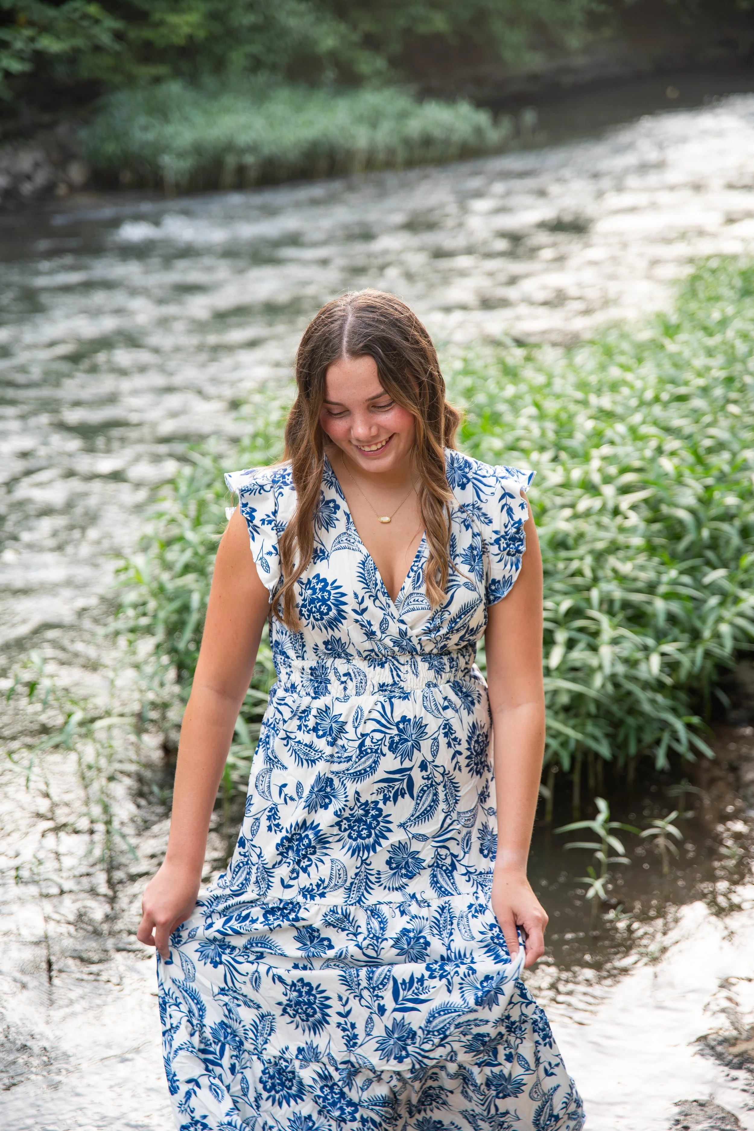 A young woman in a white and blue floral dress standing in a shallow river, smiling and looking down.
