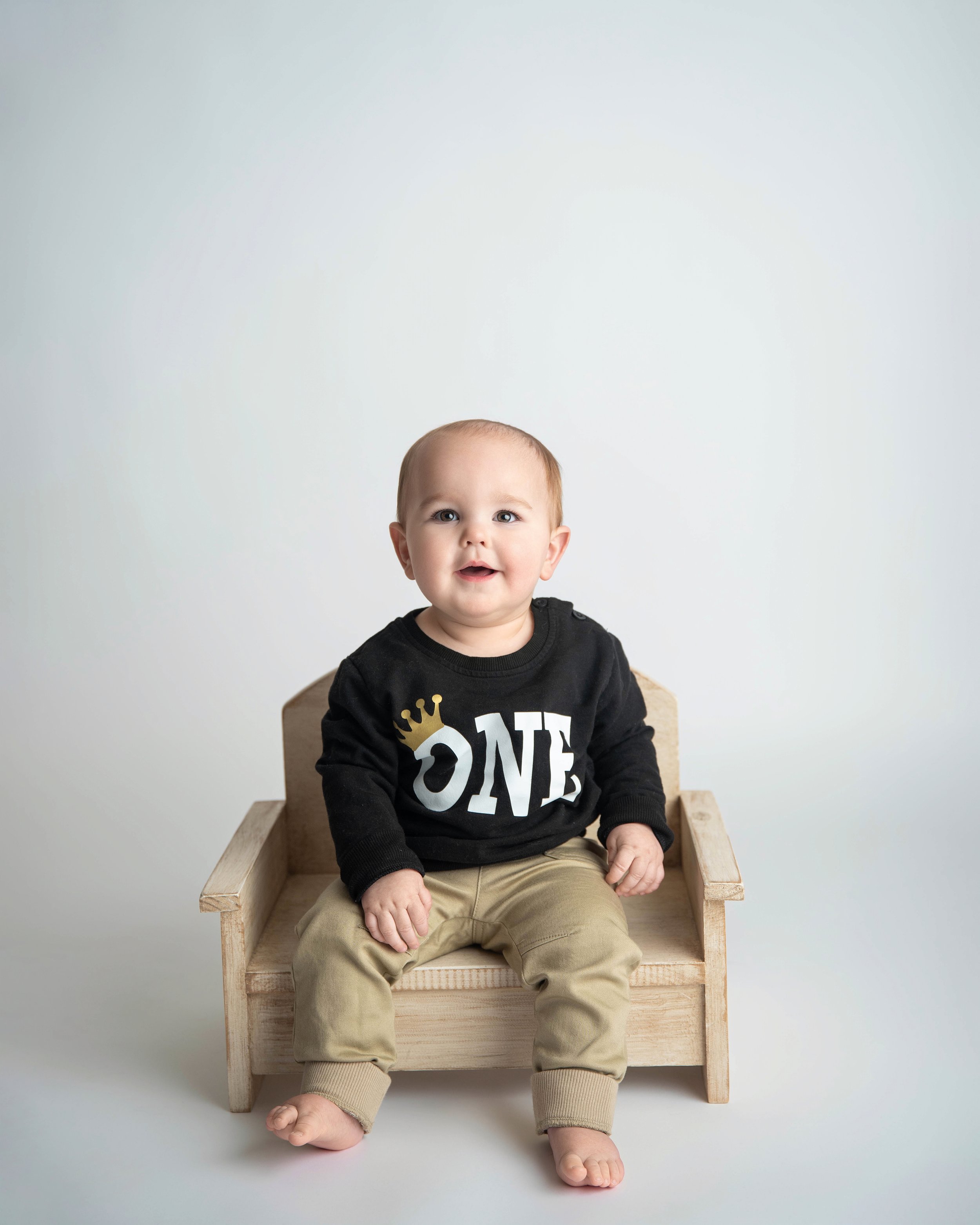 A smiling baby boy sitting on a wooden bench, wearing a black sweatshirt with 'ONE' and a gold crown logo, beige pants, and no shoes.