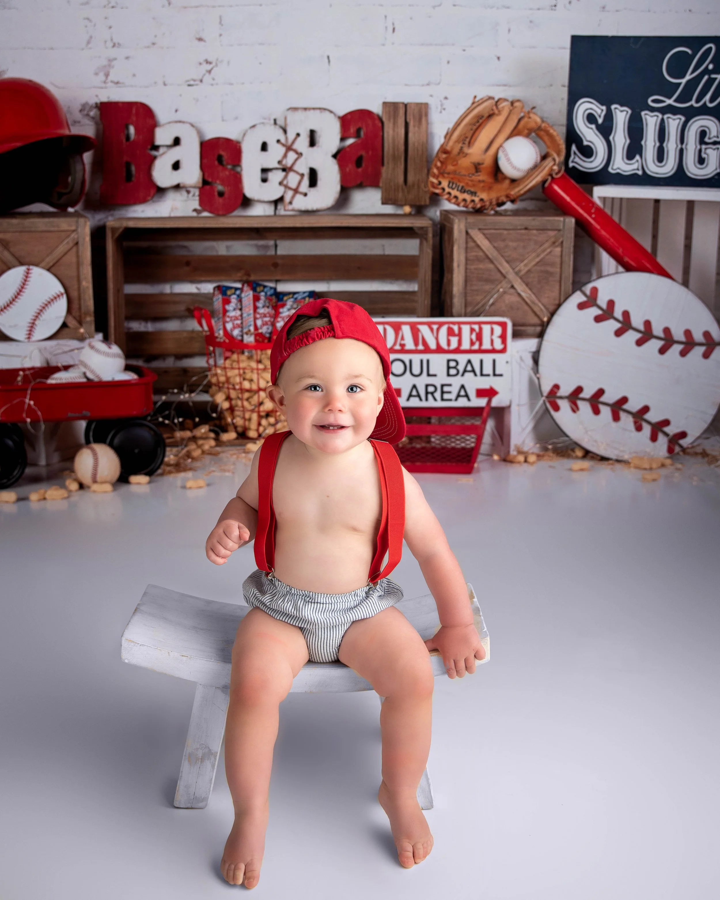 Young child with a red backward cap, red suspenders, and shorts, sitting on a small white bench in a baseball-themed indoor setting with baseballs, bats, and sports signs in the background.