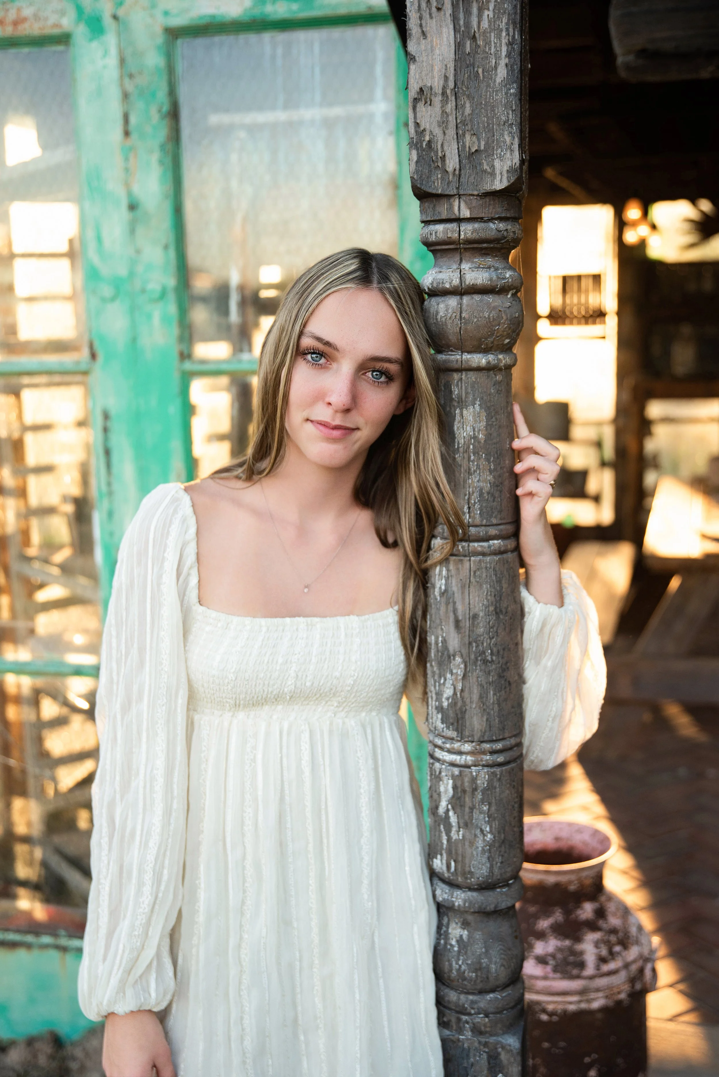 A young woman with long blonde hair and blue eyes wearing a cream-colored, long-sleeved dress standing outdoors near a weathered wooden post. She is holding onto the post with her right hand, with a rustic barn or shed in the background, bathed in wa