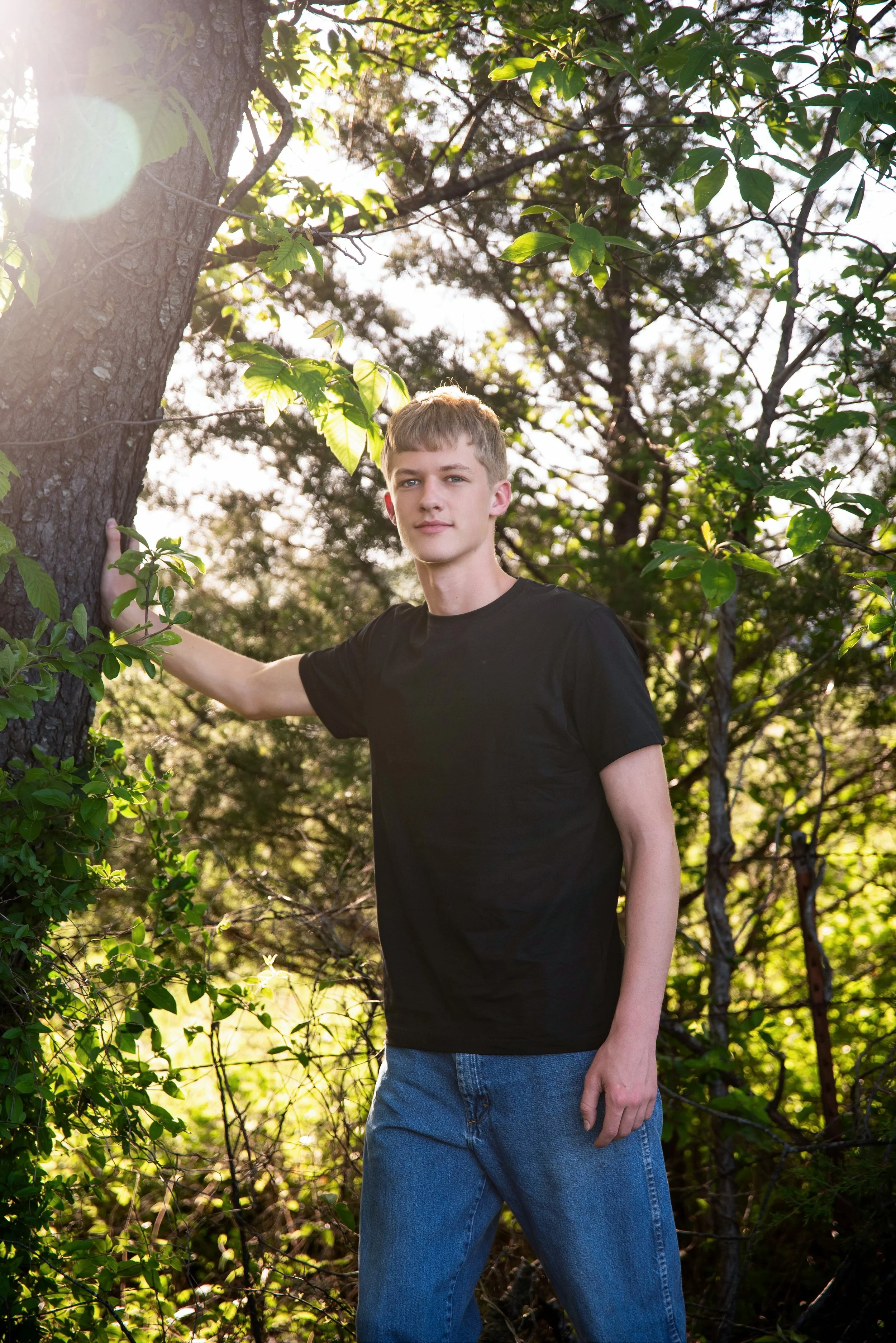 A young man wearing a black t-shirt and blue jeans standing outdoors near a tree, with sunlight filtering through the leaves.