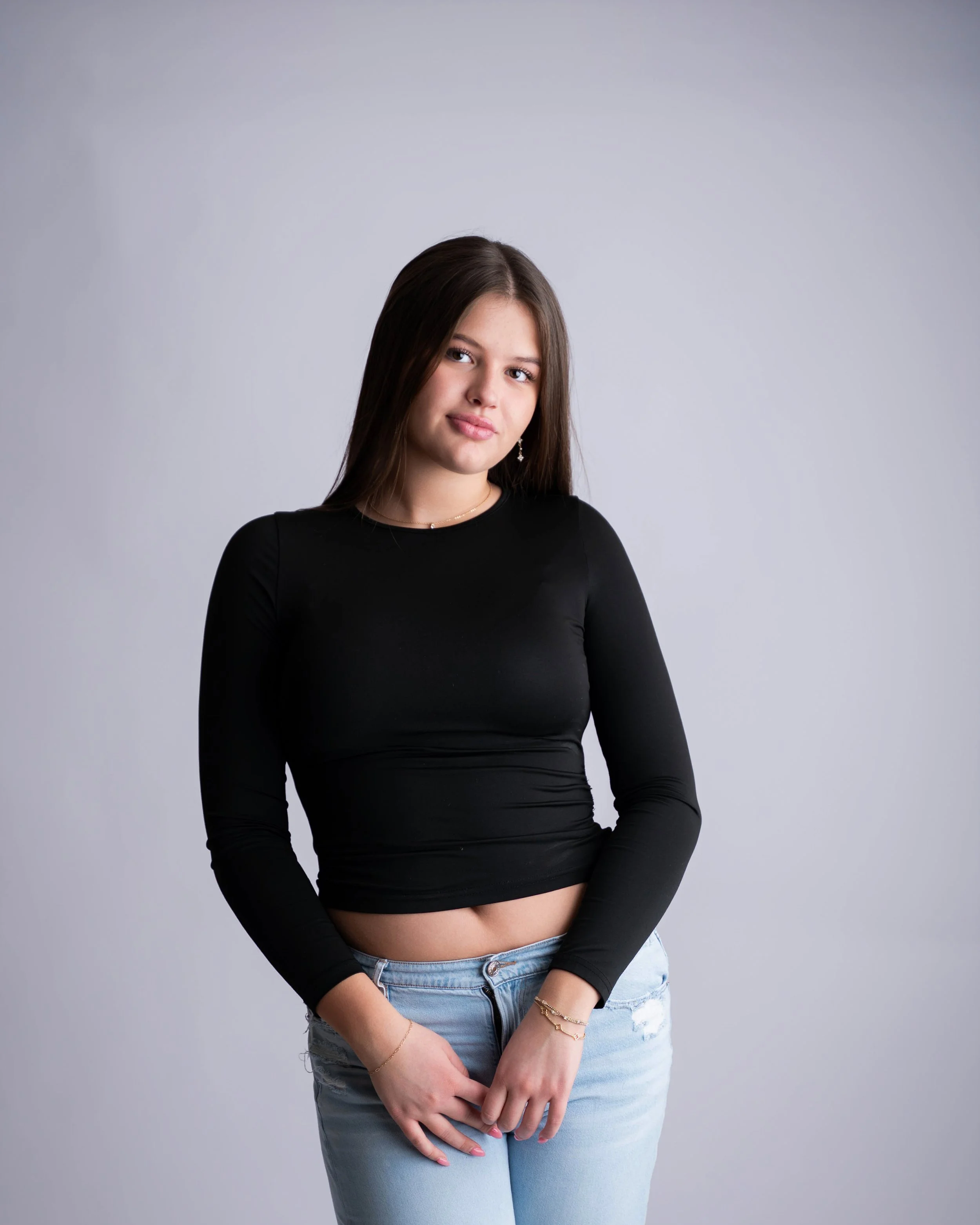 A young woman with long brown hair, wearing a black long-sleeve crop top and light blue jeans, standing against a plain gray background.