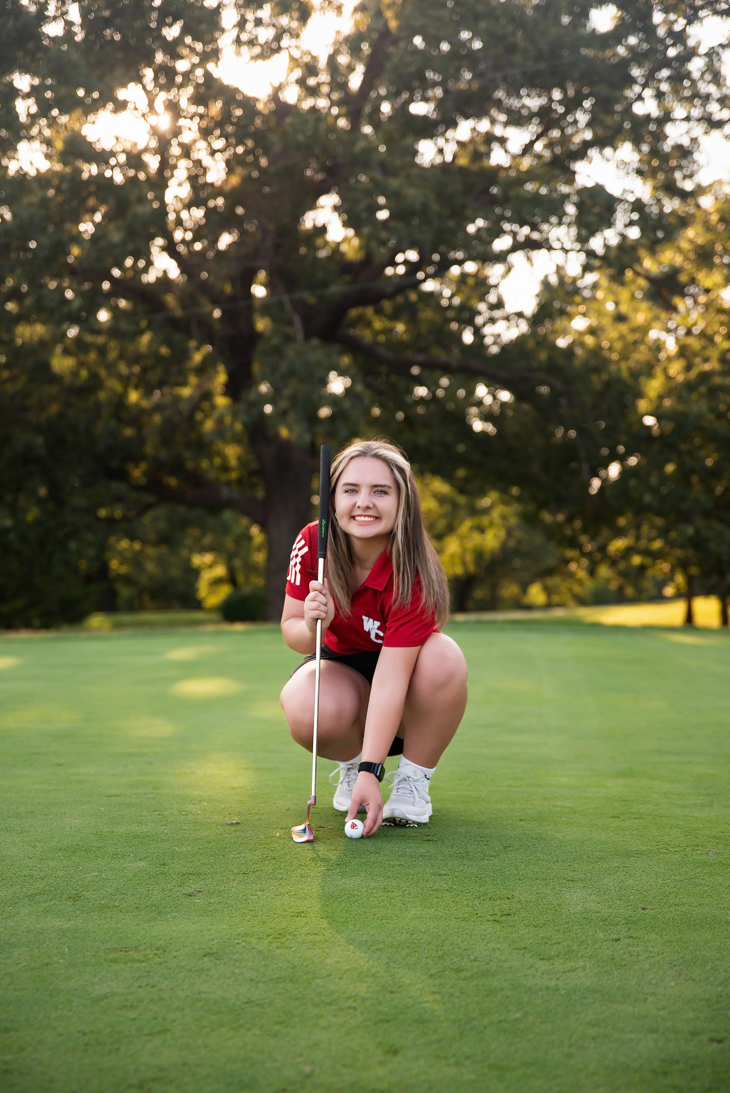 A young woman in a red sports shirt and black shorts crouches on a golf course, holding a golf club and preparing to putt. She has long hair and is smiling at the camera with trees and sunlight in the background.