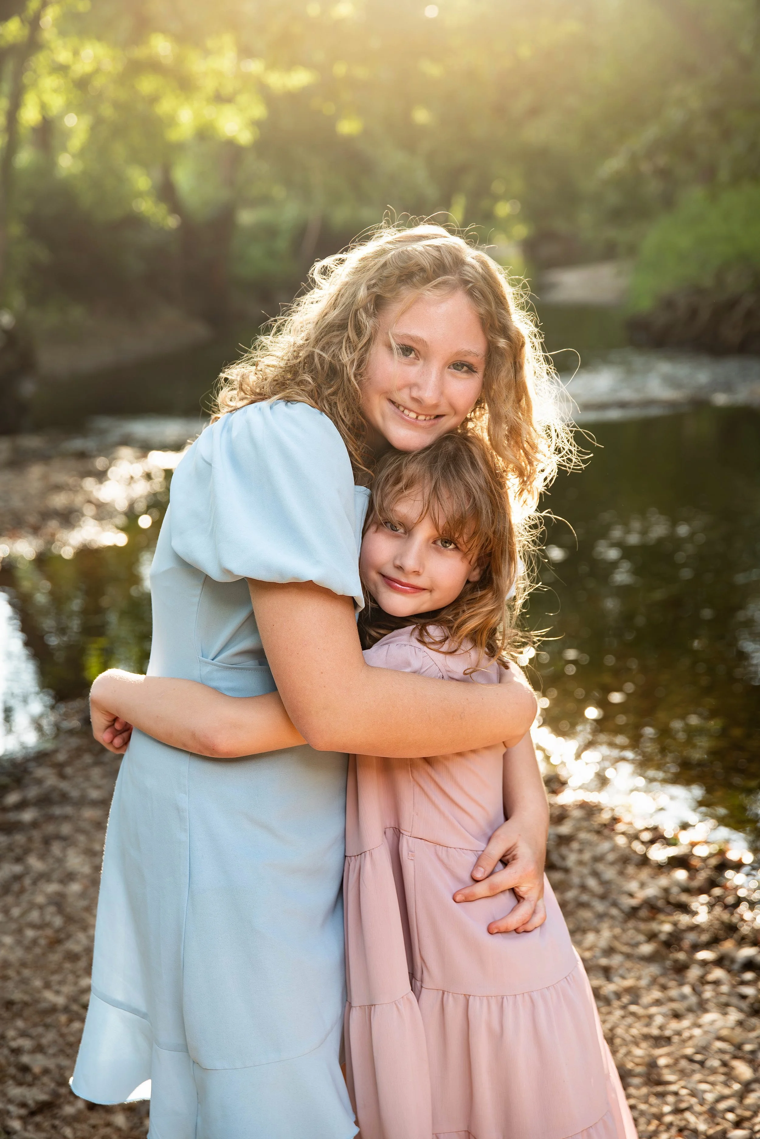 Two young girls hugging outdoors near a river, with sunlight filtering through trees in the background.