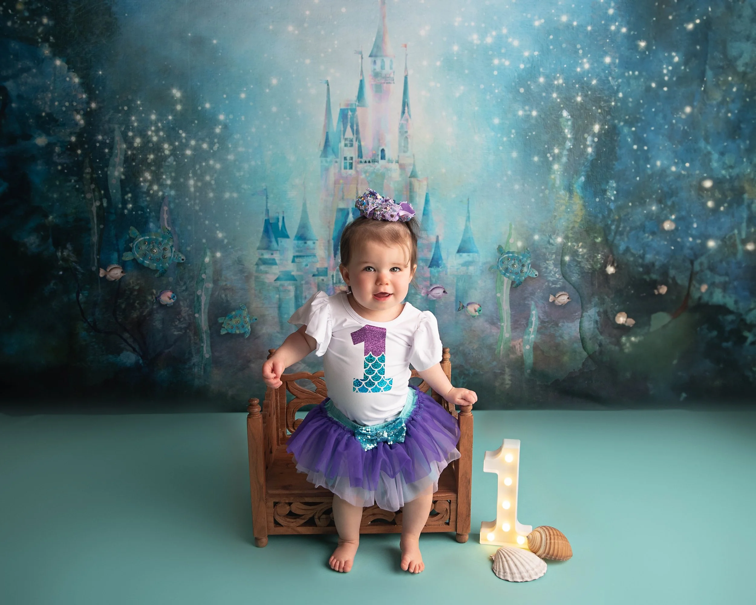 A young girl celebrating her first birthday, wearing a purple tutu and a white shirt with a large number 1 on it, standing barefoot on a light blue floor in front of an underwater and castle-themed backdrop, with seashells and a lit number 1 decorati