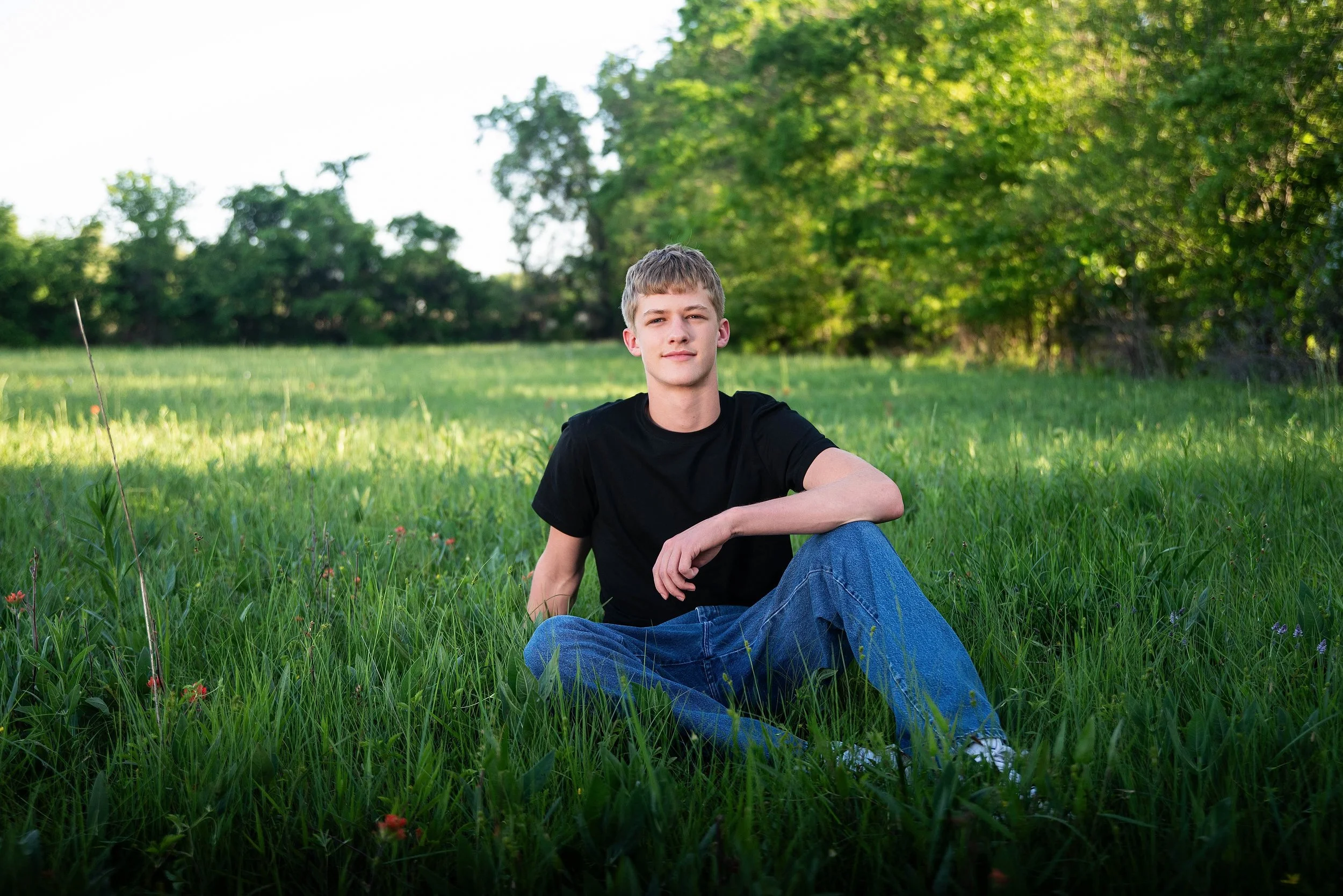 A young man sitting in a grassy field with trees in the background on a sunny day.