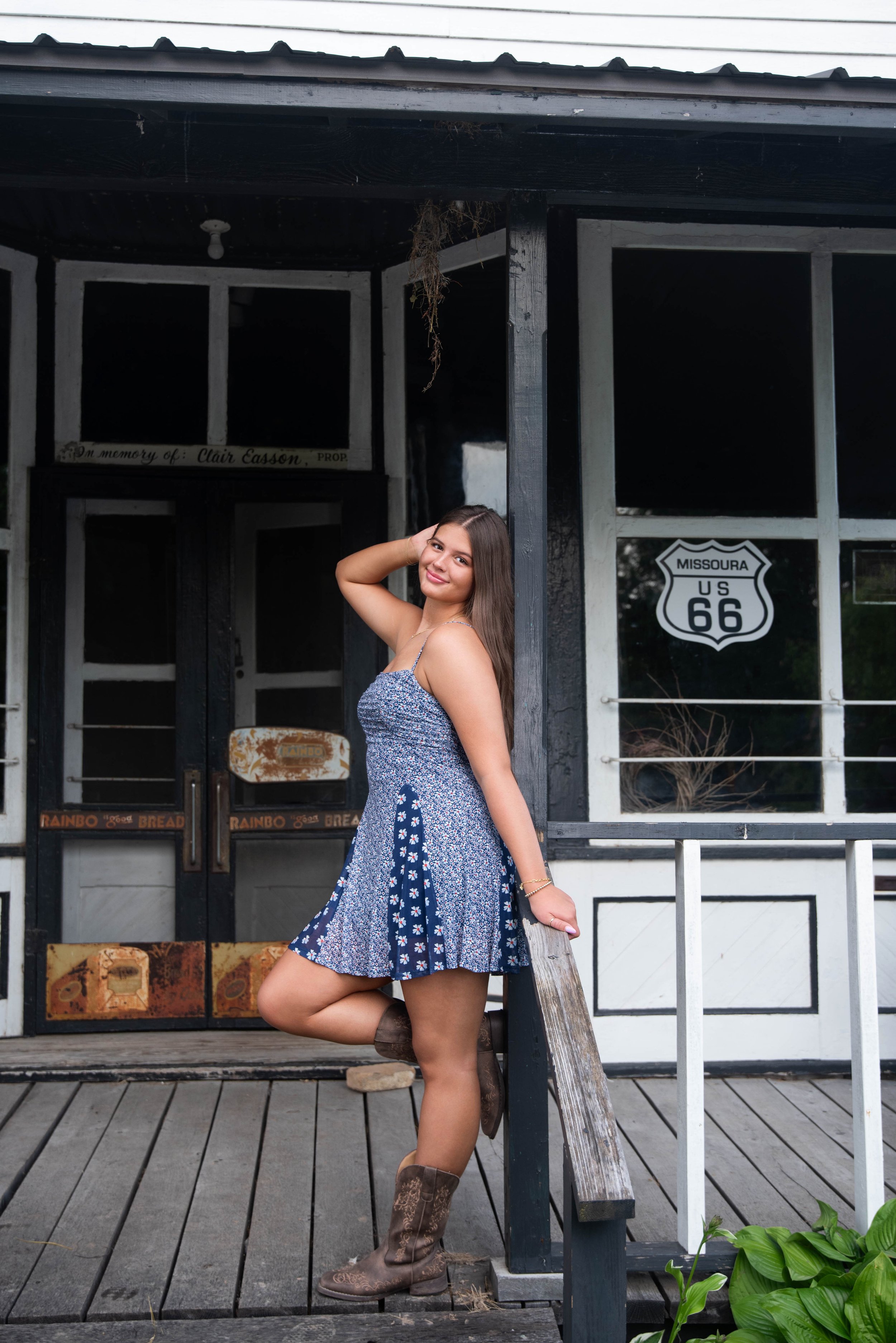 A young woman with long brown hair wearing a blue floral dress and cowboy boots, posing with one foot on the step in front of a rustic storefront with a Missouri US Route 66 sign.