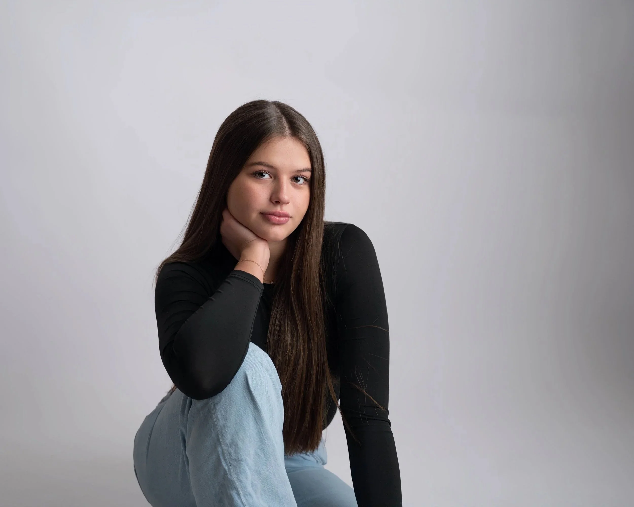A young woman with long brown hair and fair skin, wearing a black long-sleeve top and light blue jeans, sitting against a plain light gray background.