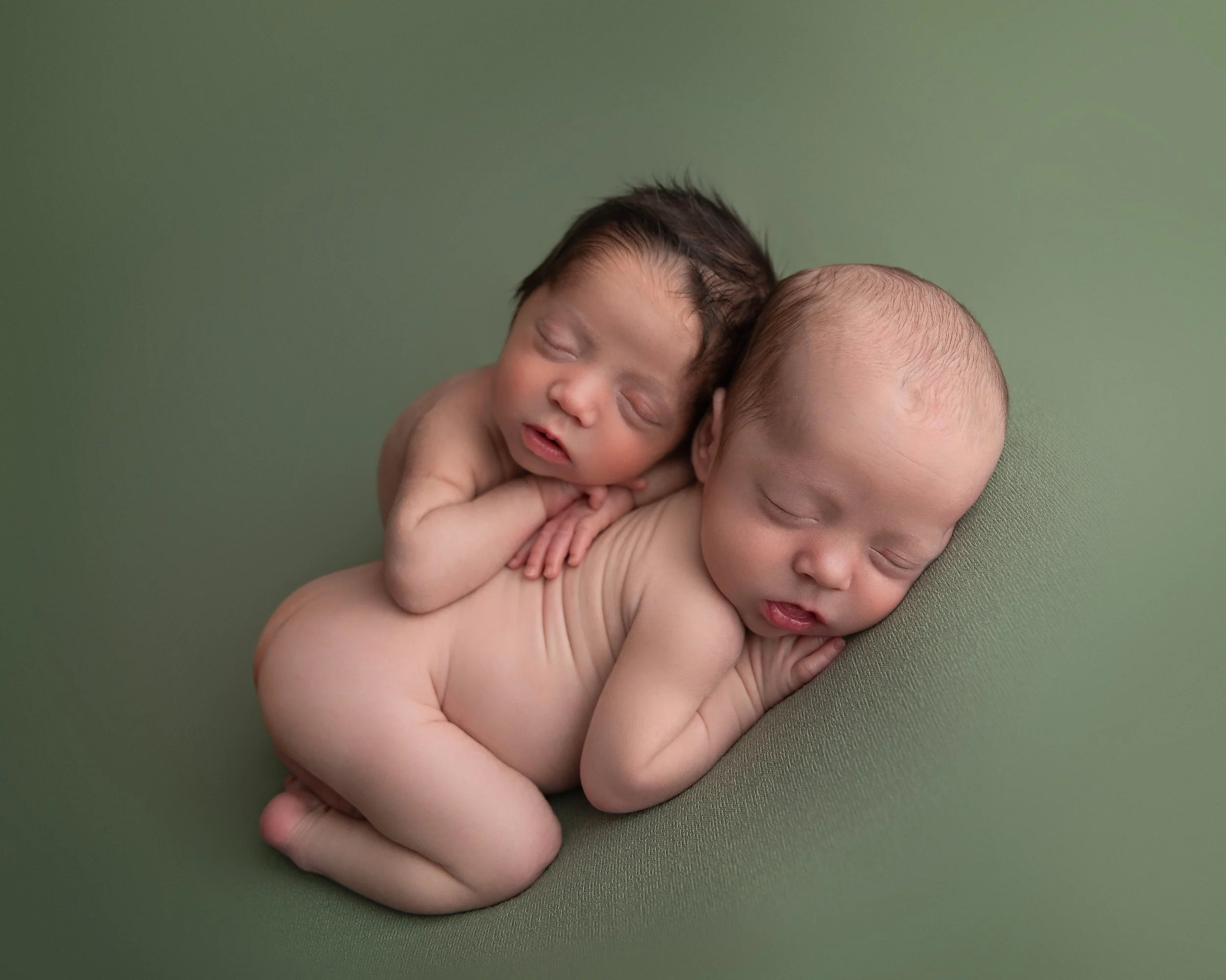 Two newborn babies sleeping, one with dark hair and one with light hair, lying on a green surface.