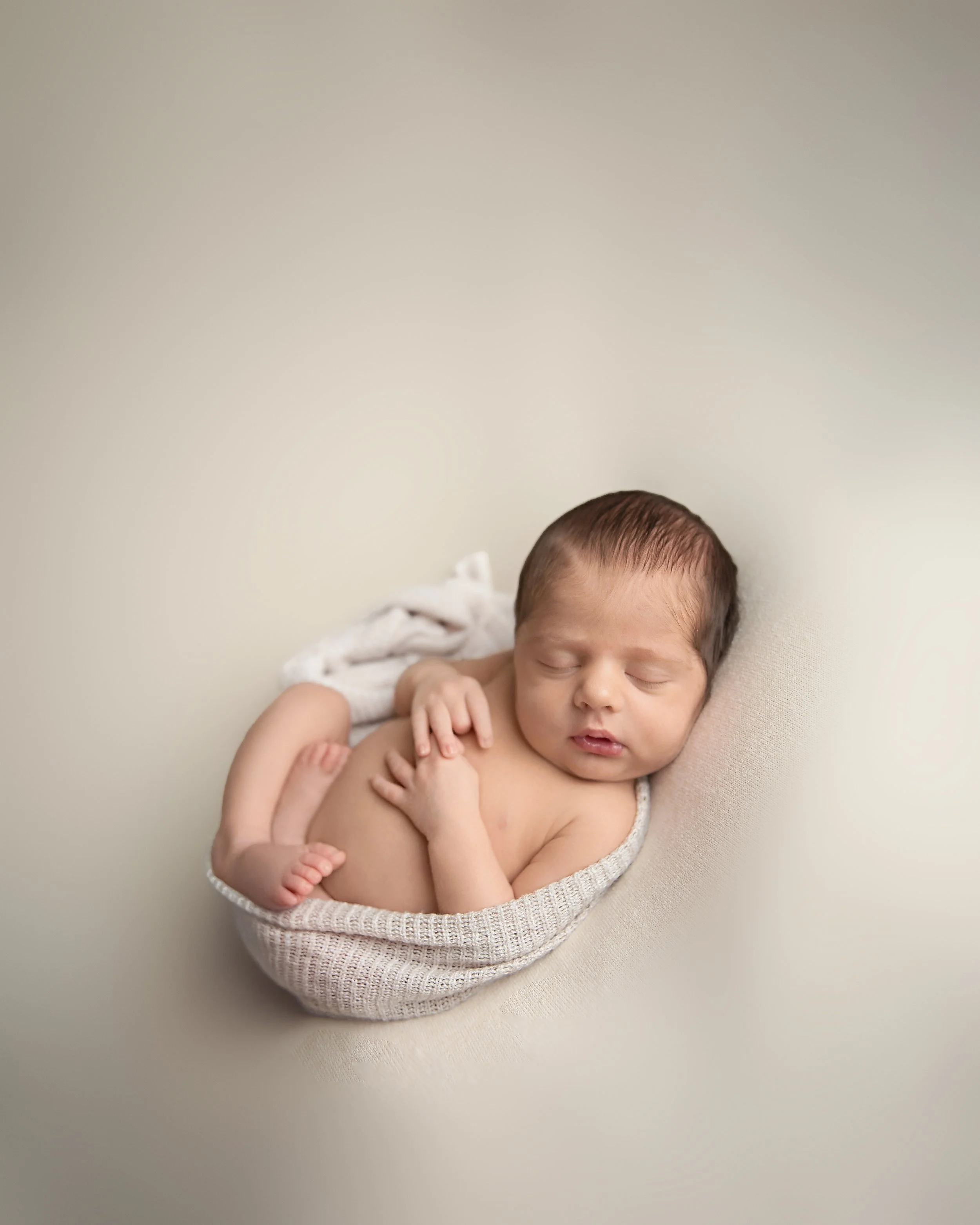 A sleeping newborn baby curled up in a knitted wrap on a light-colored surface