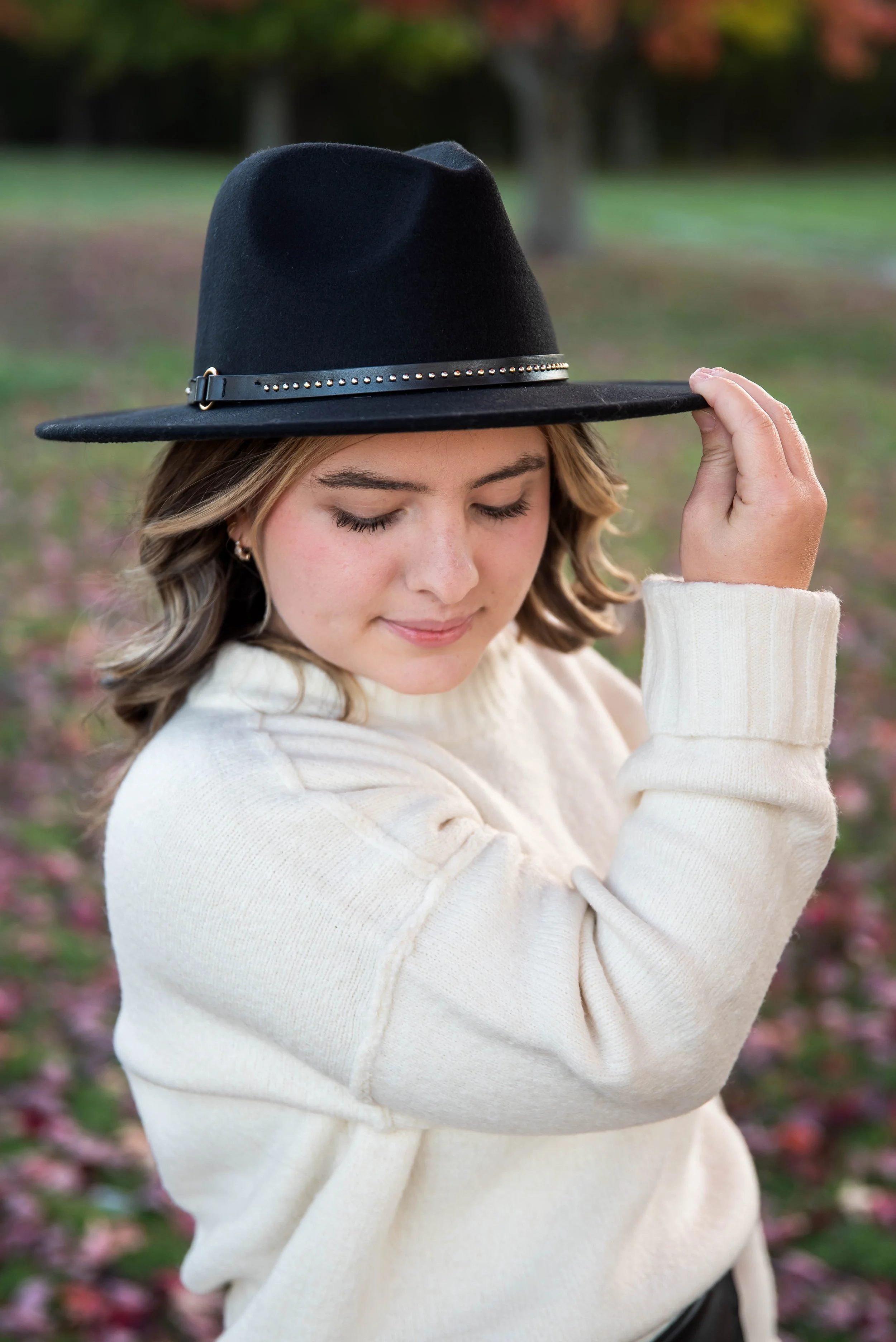A young woman with light brown hair wearing a black wide-brimmed hat with silver accents, a cream-colored sweater, and earrings, standing outdoors in a park with autumn foliage in the background, eyes closed and smiling gently.
