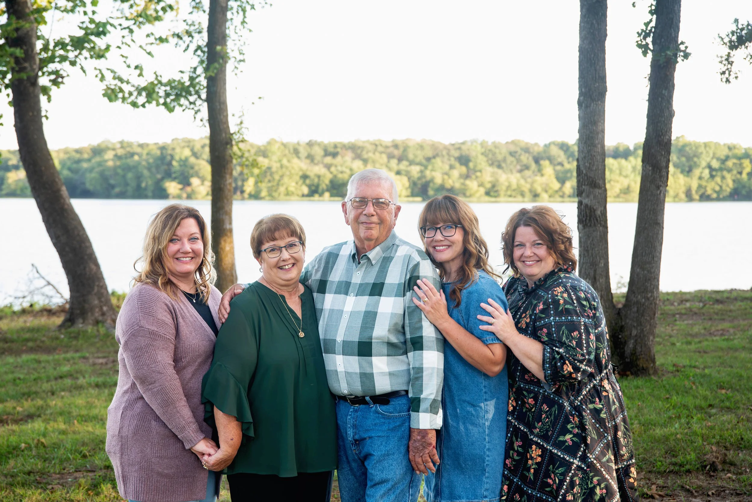 Five people standing outdoors near a lake, smiling, with trees in the background.