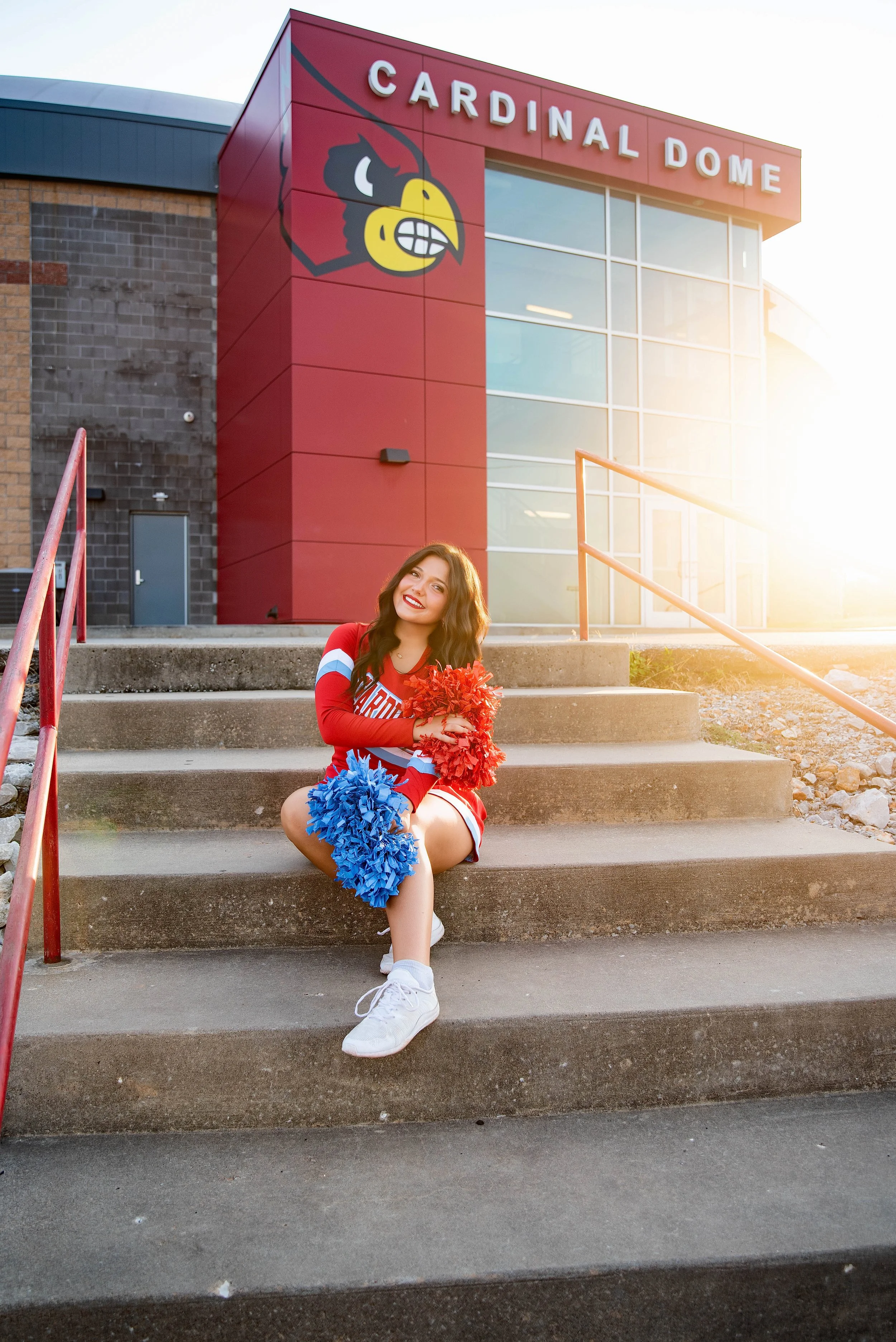 A cheerleader sitting on concrete steps outside the Cardinal Dome stadium, holding red and blue pom-poms, with the stadium's sign and logo visible in the background during sunset.