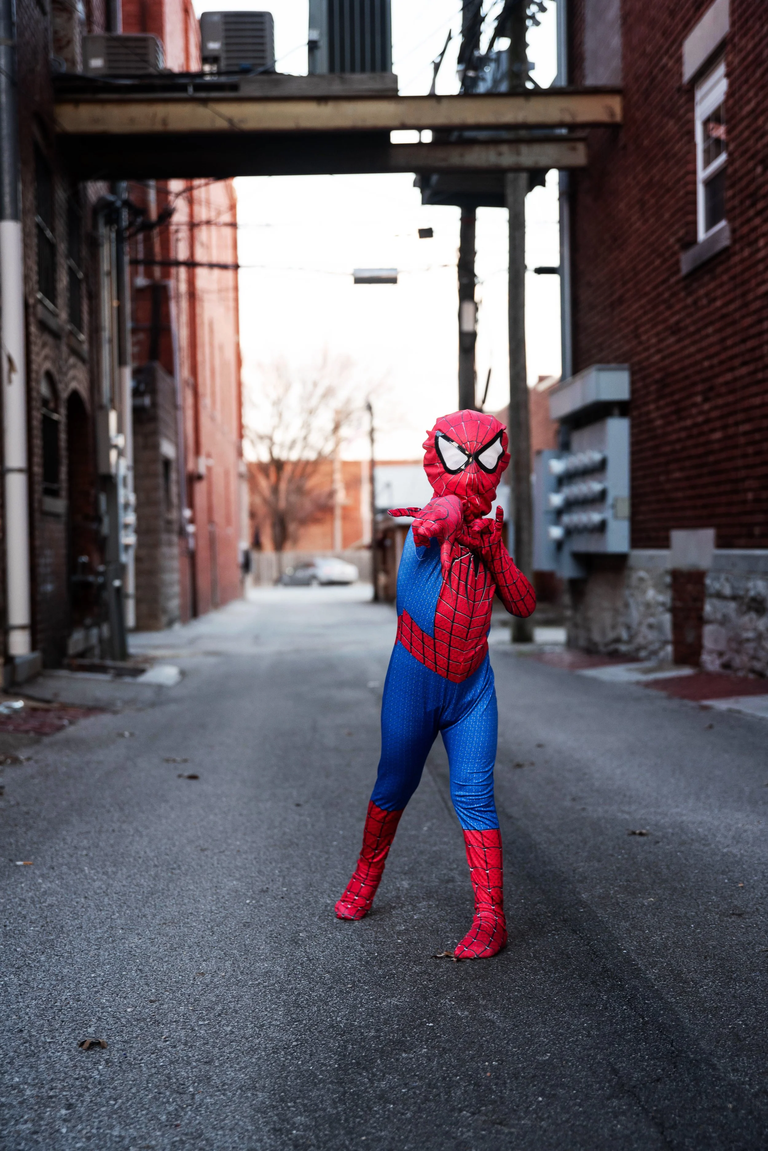 Child dressed as Spider-Man in a city alley pointing towards the camera.